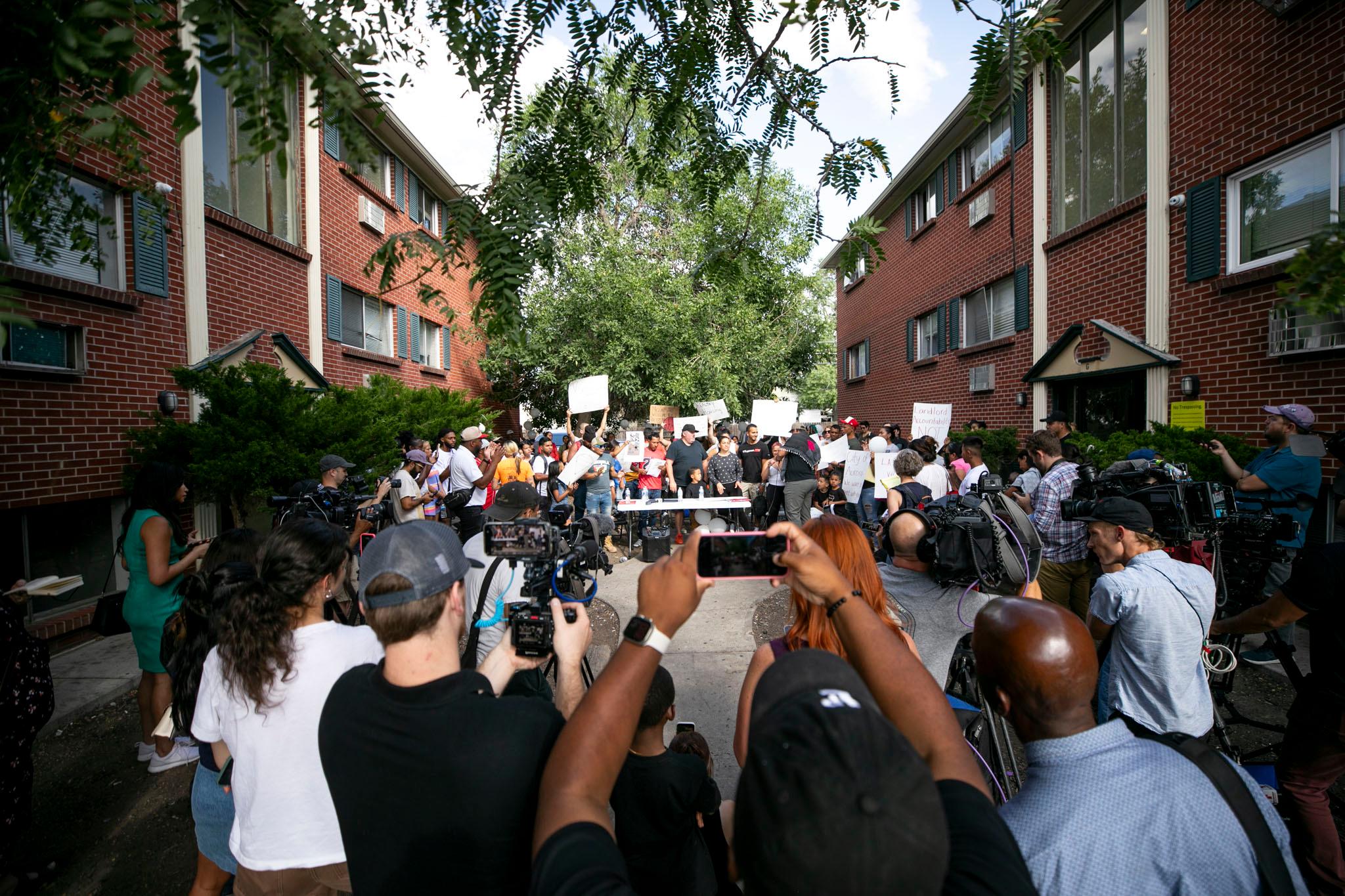 Dozens of people are gathered beneath a brick apartment building. Some hold signs above their heads. People in the foreground point cameras towards the larger group.