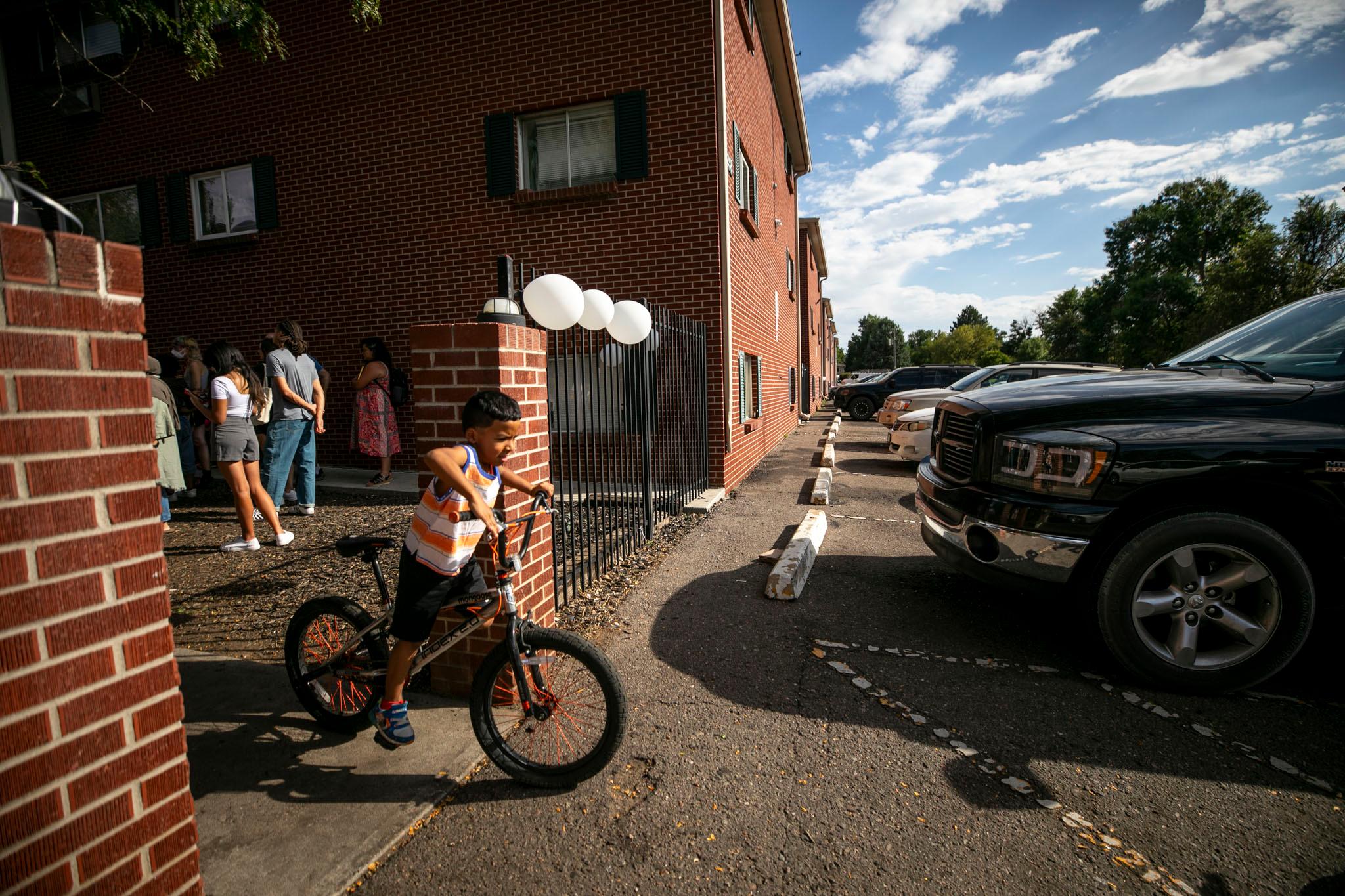 A kid on a bike rides out of the courtyard of a row of brick buildings. Cars are parked in spaces on the right.