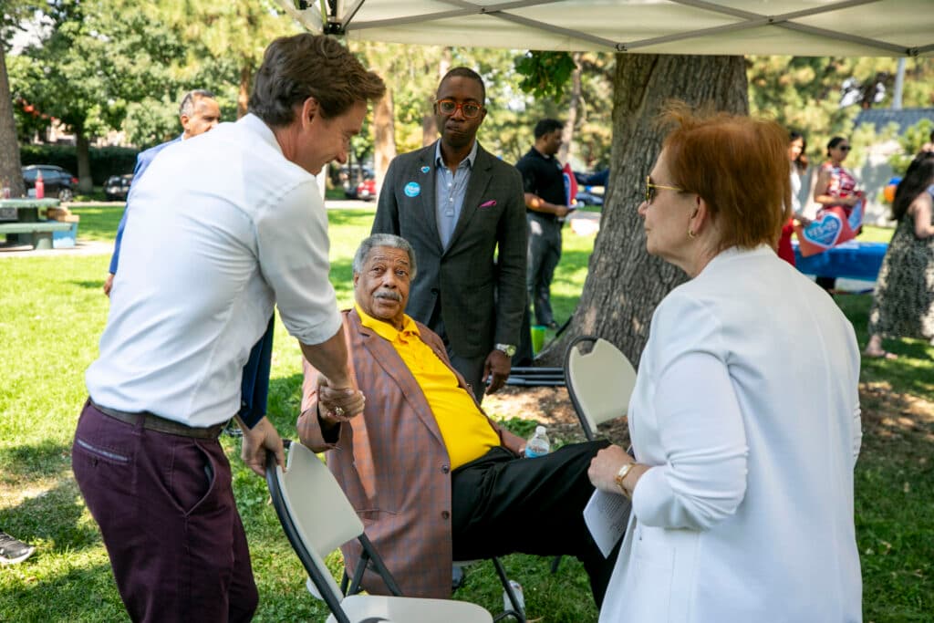 A man with grey hair and a grey caterpillar mustache, sitting in a chair, looks at a man in a dress shirt and tie as they shake hands. A few people watch.