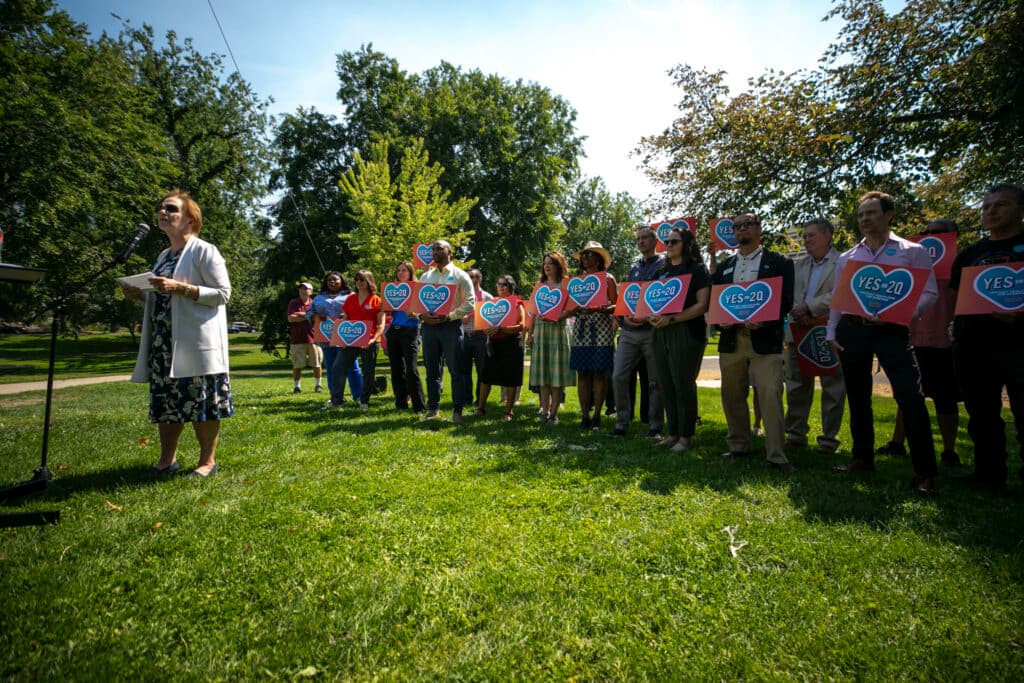 A woman in a white sweater over a blue dress, and in dark shades, speaks at a microphone on a green lawn, in front of a line of people holding signs with hearts on them that say &quot;YES ON 2Q.&quot;