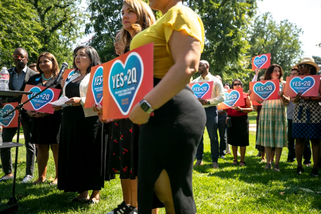 A woman in a black dress on the left side of the frame speaks into a microphone. She's in one line of people holding signs with hearts on them that say &quot;YES ON 2Q;&quot; there's another line behind them.