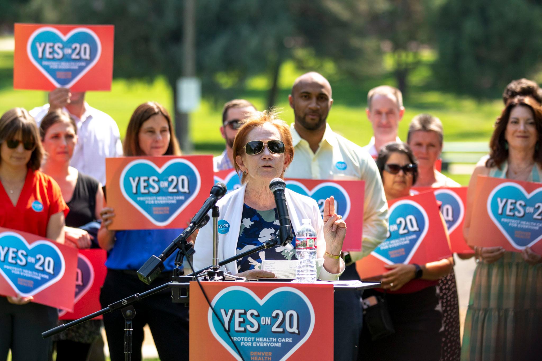 A woman in a white sweater over a blue dress, and in dark shades, speaks at a microphone on a green lawn, in front of a line of people holding signs with hearts on them that say "YES ON 2Q."