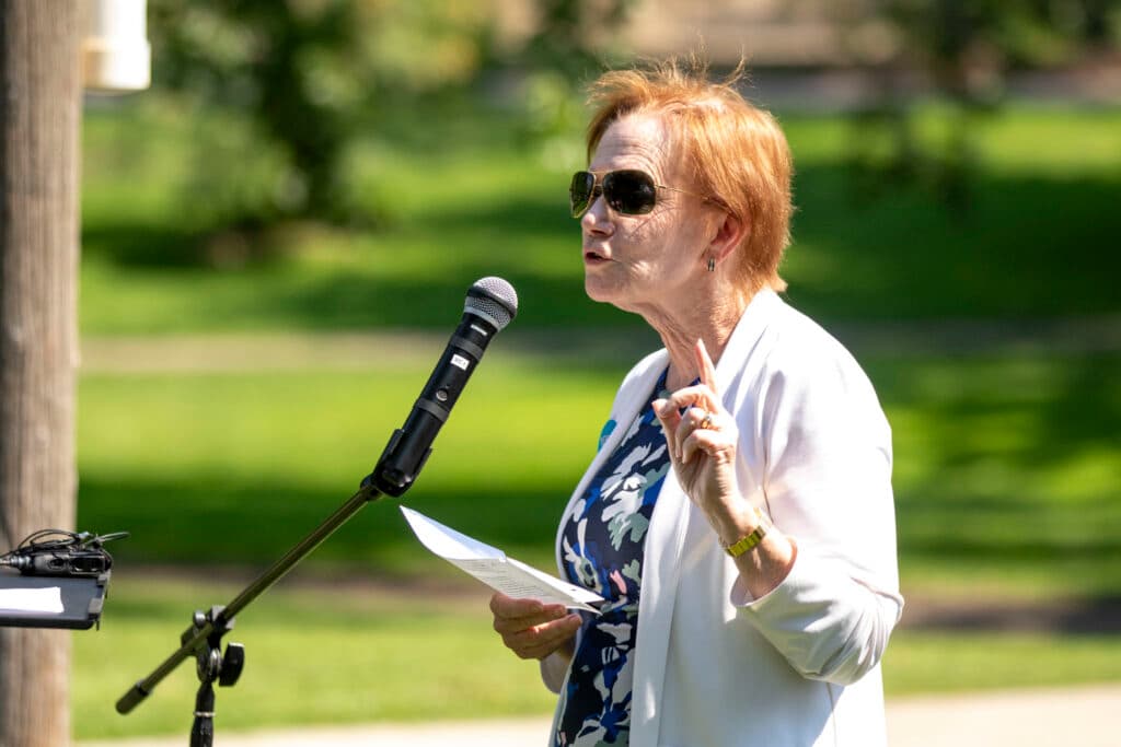 A woman in a white sweater over a blue dress, and in dark shades, speaks at a microphone on a green lawn. She's waving her hand as if she's making a point.