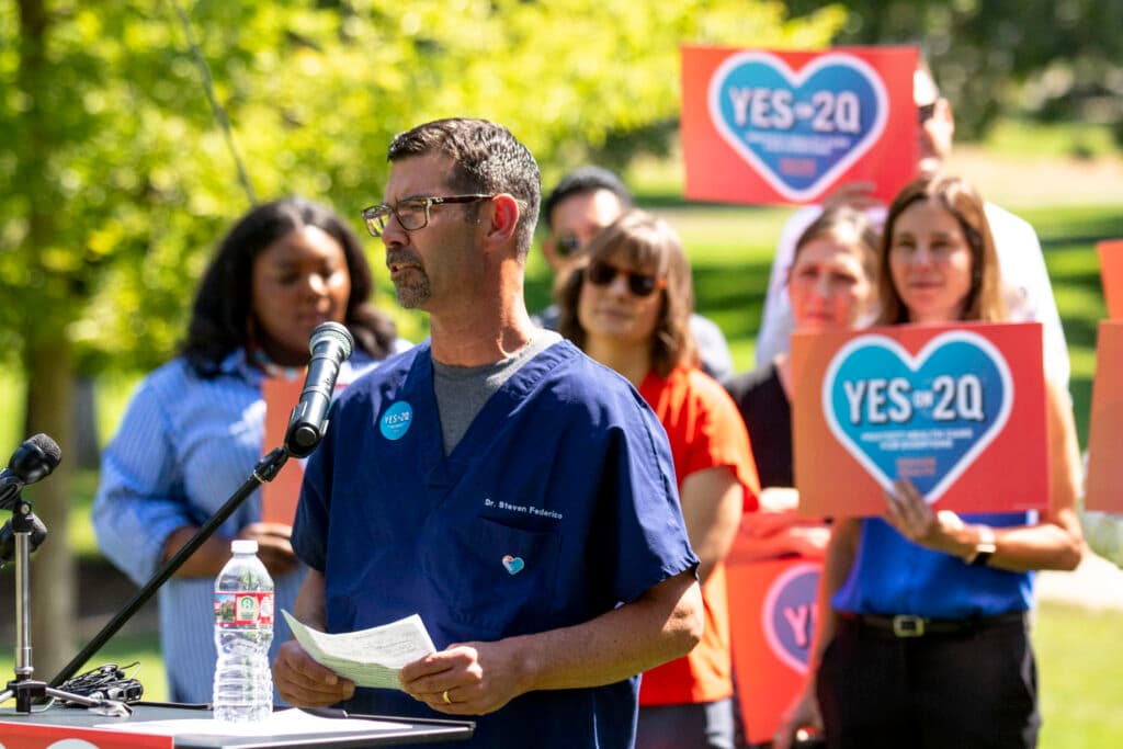 A man in blue scrubs speaks at a microphone, on a green lawn, in front of a line of people holding signs with hearts on them that say &quot;YES ON 2Q.&quot;