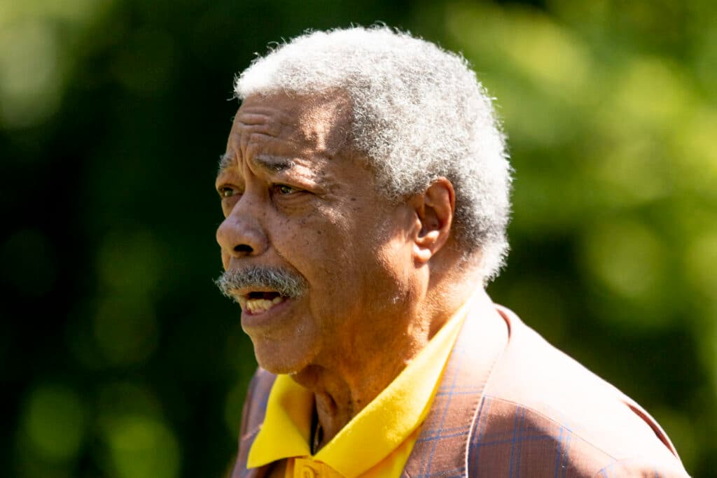 A man with grey hair and a grey caterpillar mustache speaks, presumably at a microphone, over a background of blurred green. Light glints in his eyes.