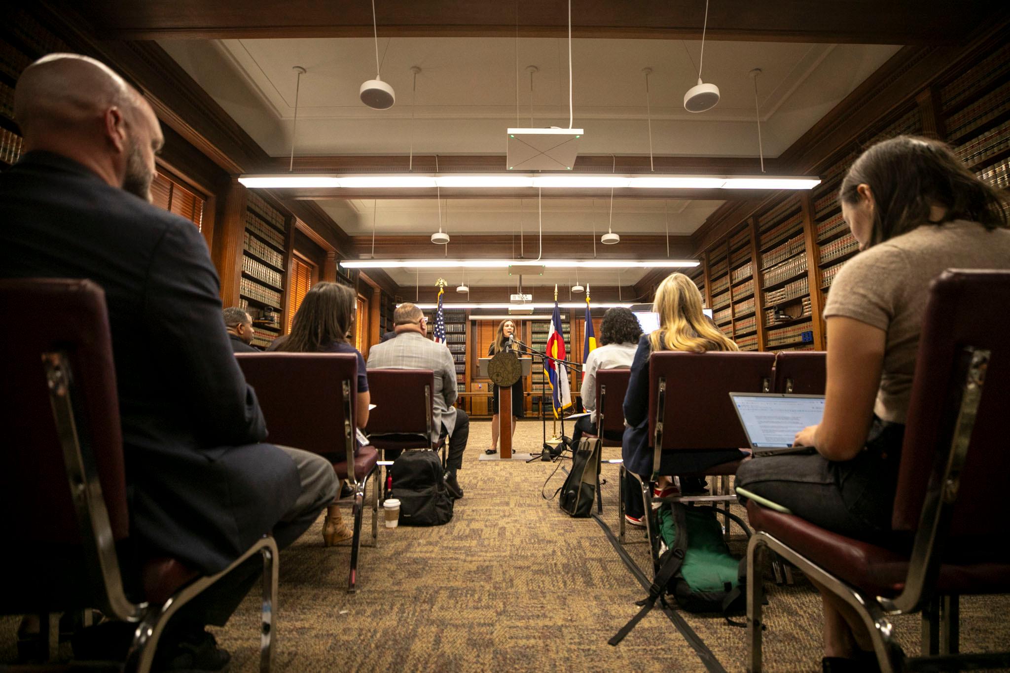 A woman in a black dress speaks at a podium before a room full of people watching.
