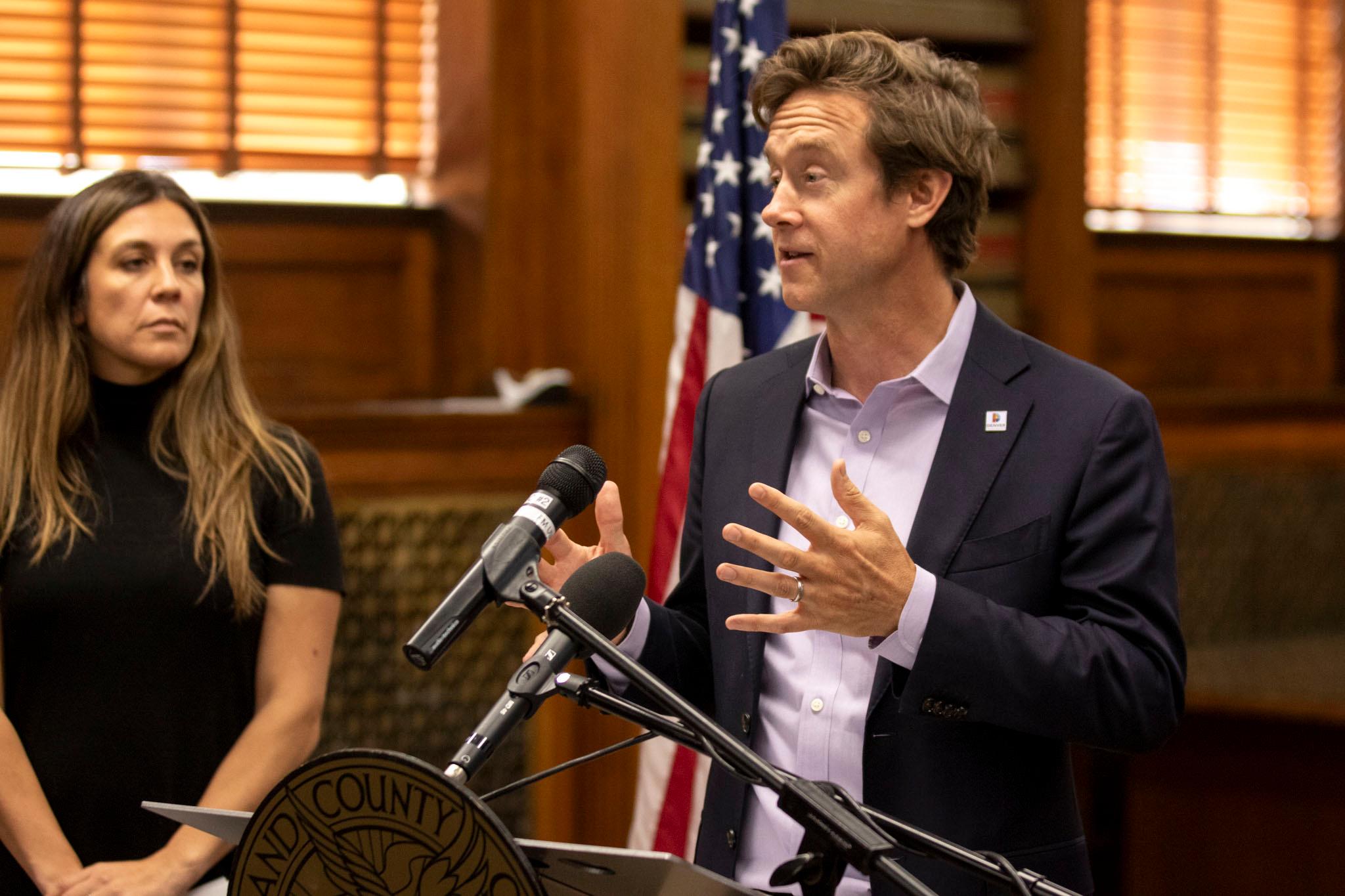 A man in a suit raises his hands as he speaks at a microphone; an American flag and a woman in black stand behind him.