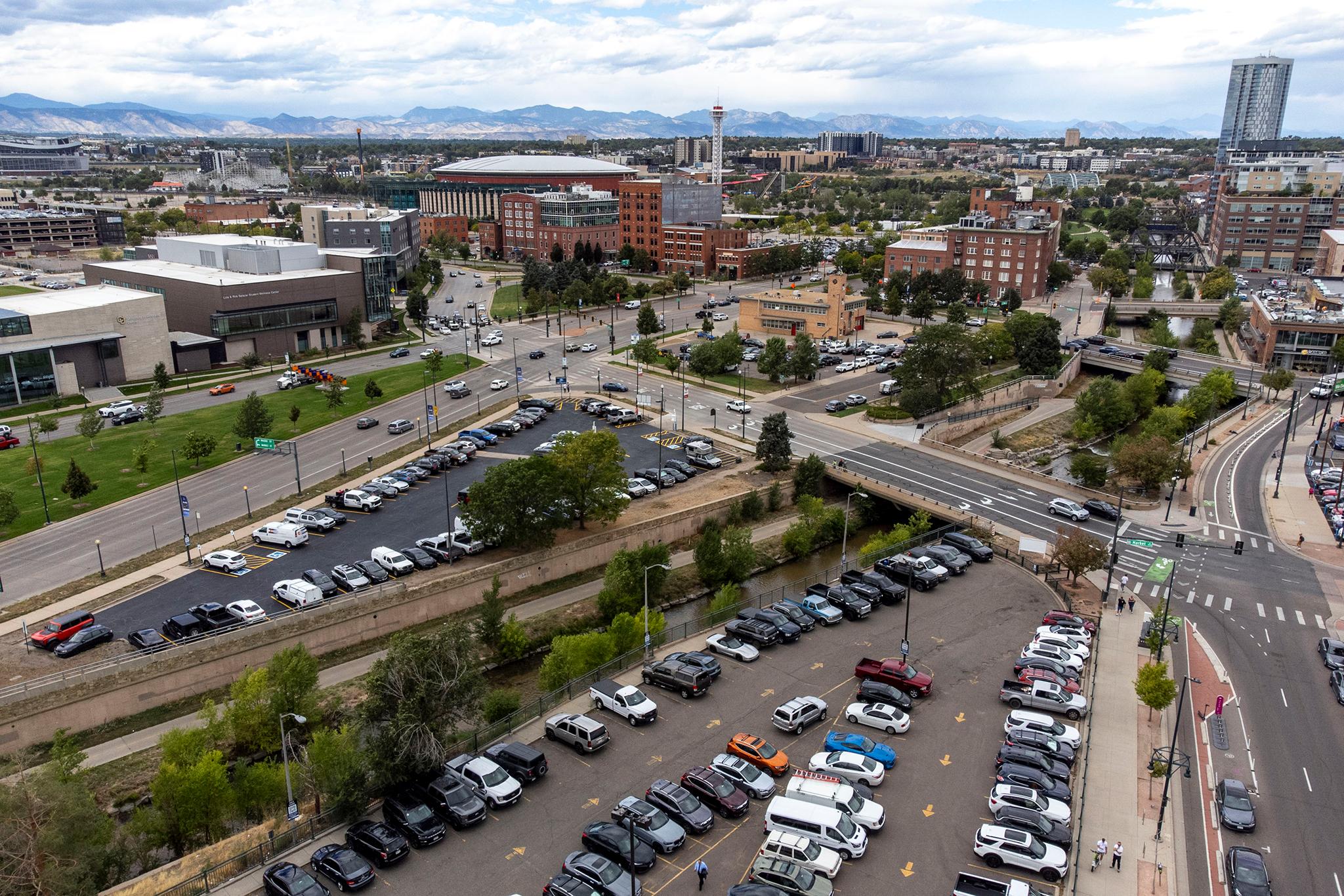 Ball Arena can be seen in context with Speer Boulevard and Market Street from an aerial view; mountains rise in the distance; cars are parked in lots at the bottom of the frame.