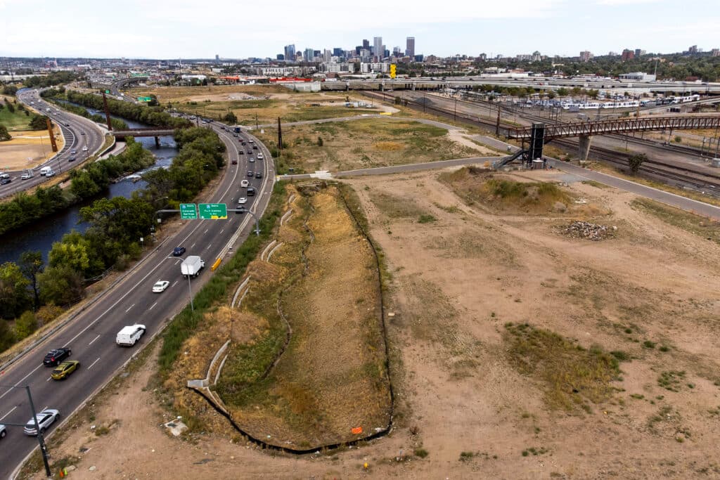 An aerial view of a large empty lot. A river and a major street run along its right side; the city's skyline rises in the distance.