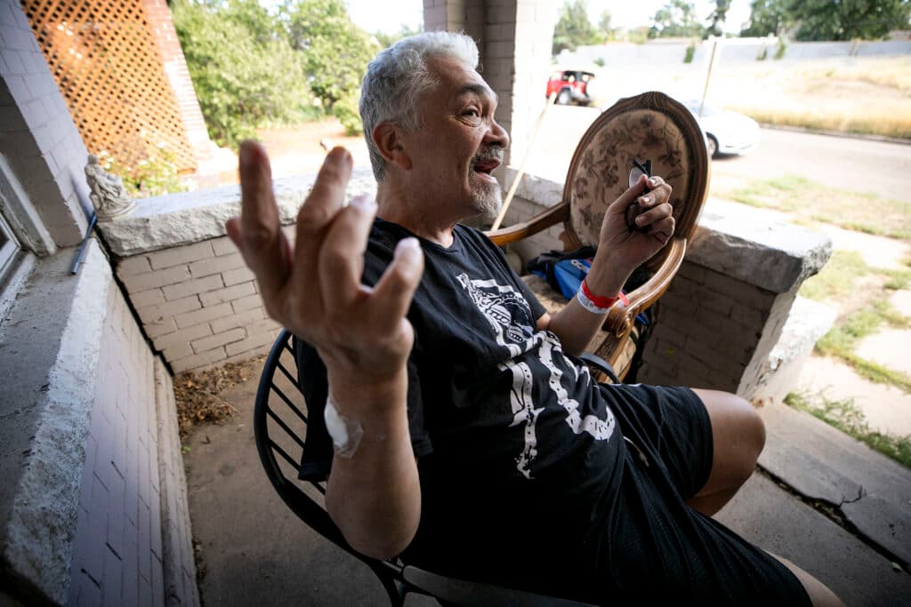 A man in a black t-shirt raises his hands as he speaks passionately, from a chair on a brick and concrete porch.