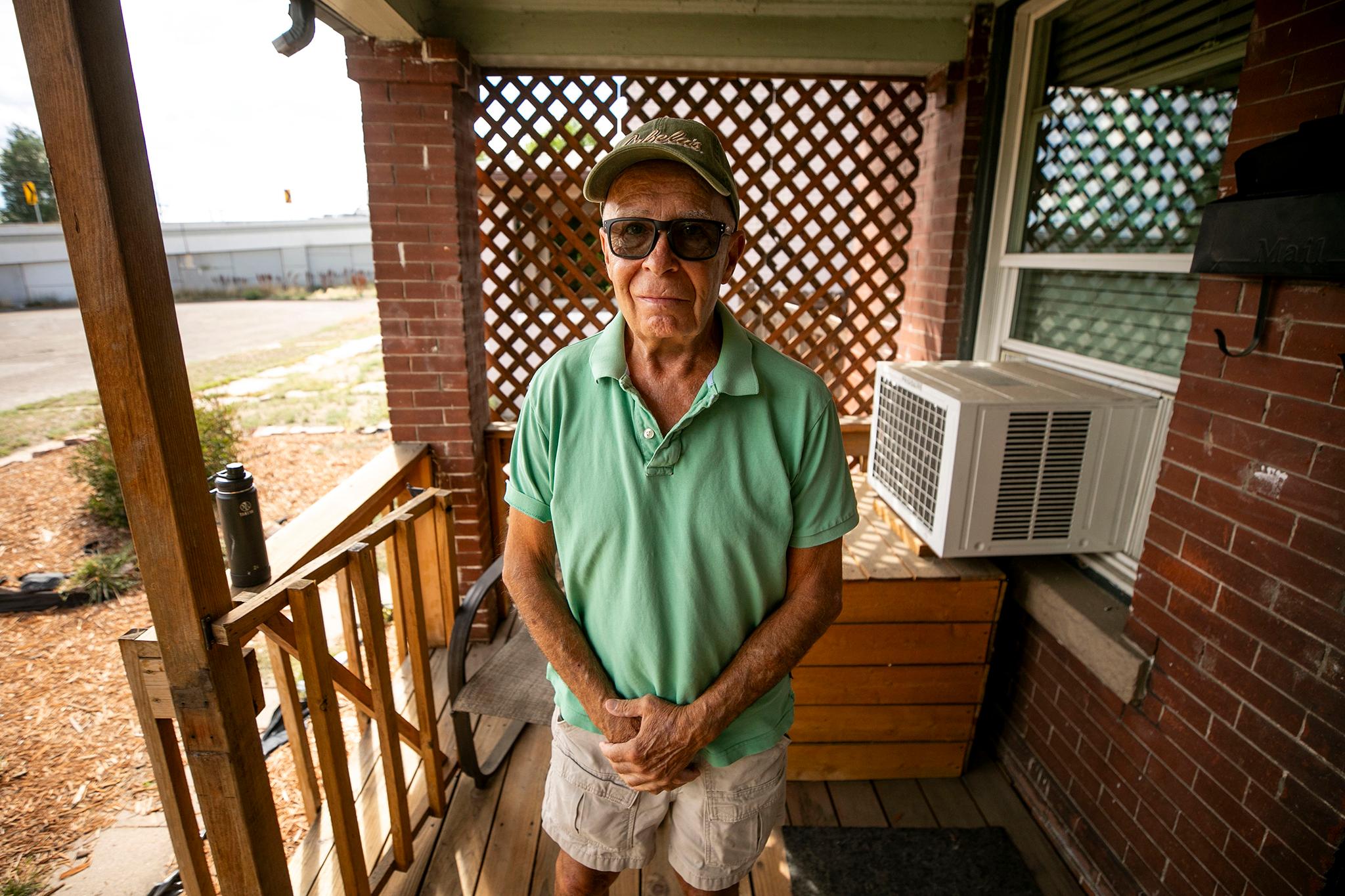 A man in a green polo and shorts stands under a porch, holding his hands in front of him and staring right into the camera.