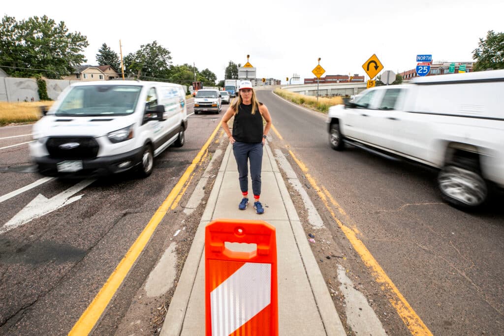 A woman in a ball cap and a black shirt stands on a thin strip of concrtete between an onramp and an offramp as trucks pass her going both ways, blurred in their motion.