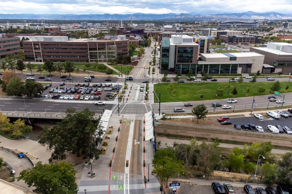 An aerial view of Larimer Street stretching towards the mountains, intersected by Speer Boulevard. Mountains rise in the distance; buildings and green grass surround the intersection.