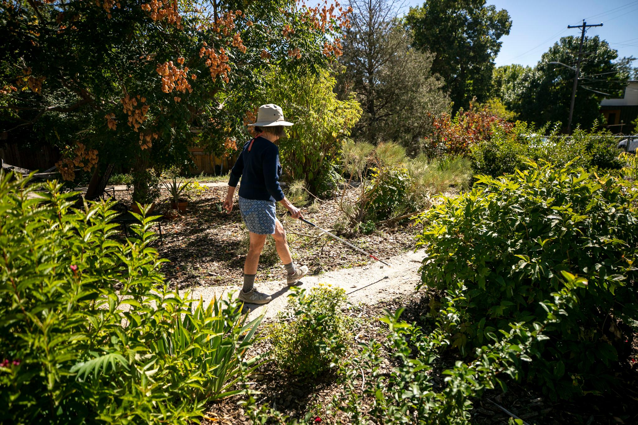 A woman in a sun hat strolls across a dirt path surrounded by green bushes and trees. She holds a white stick, which is pointed out at the ground in front of her.