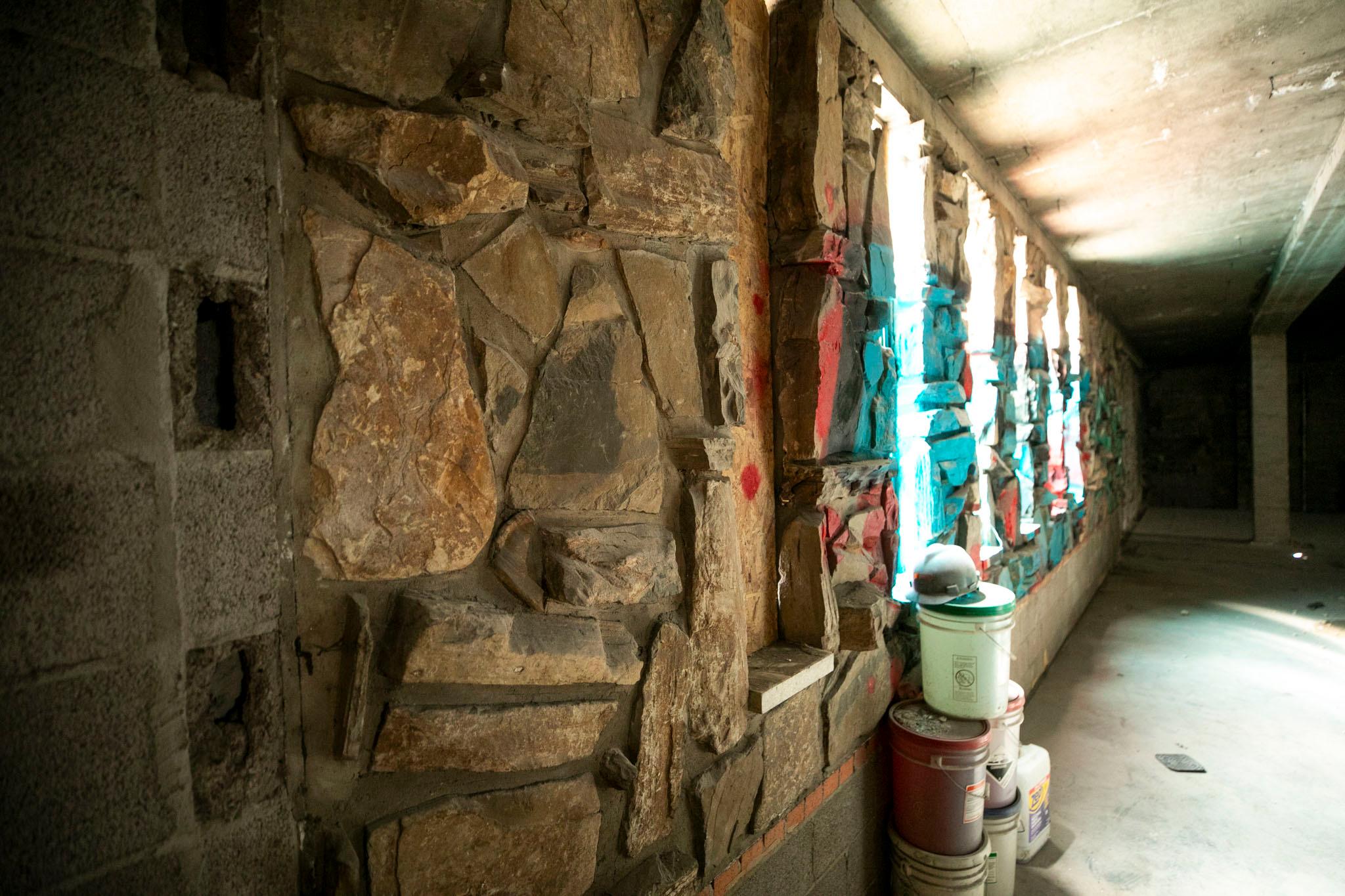 Rocks are cemented into a wall in a room that's otherwise an empty concrete shell. Sunlight pours in from windows in the background, and paint buckets and a hardhat rest beneath them.