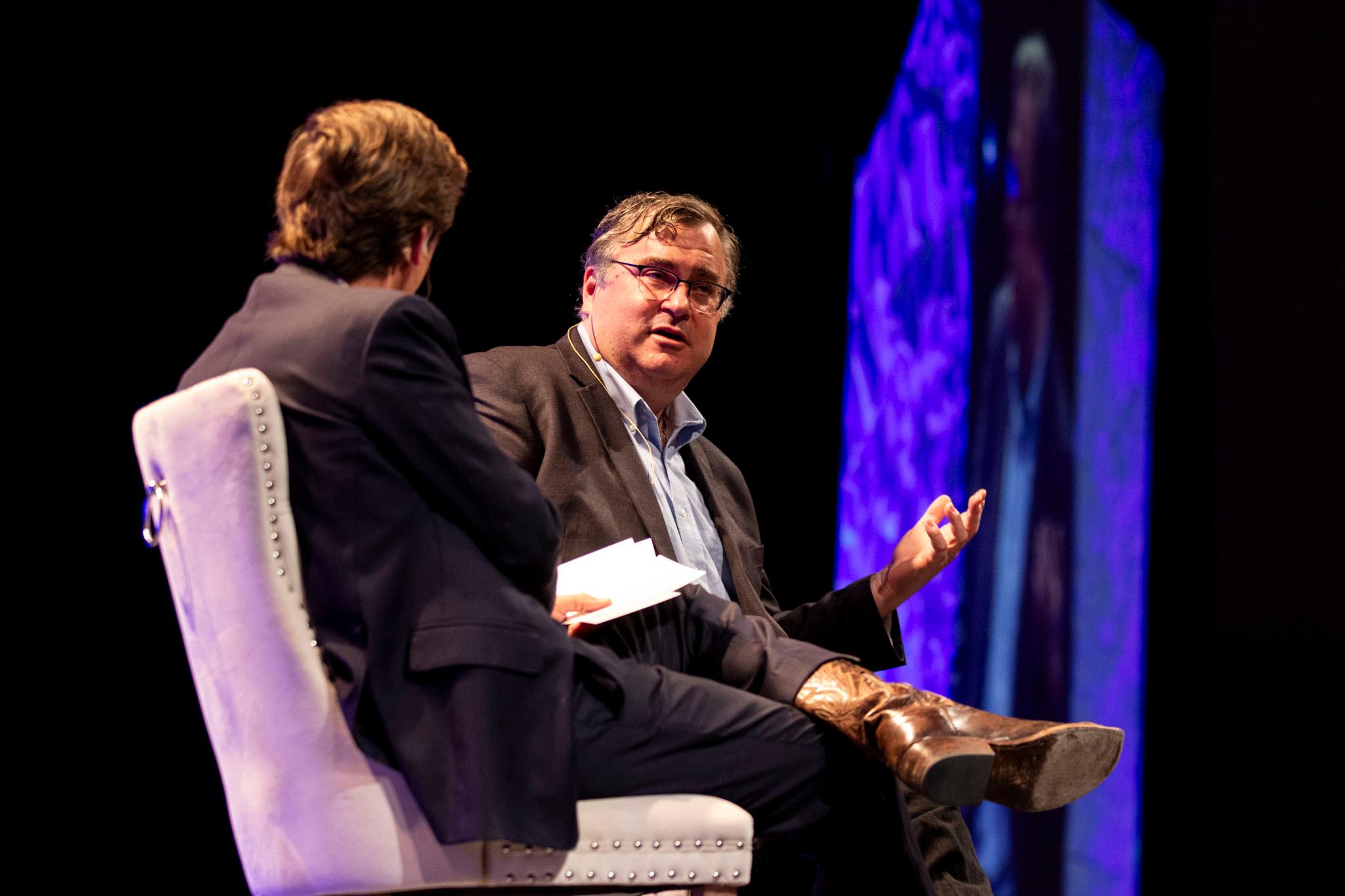 Teo men in suits sit on a stage, speaking in white plush chairs; the angle is over the shoulder of one of them; the other talks with his hands.