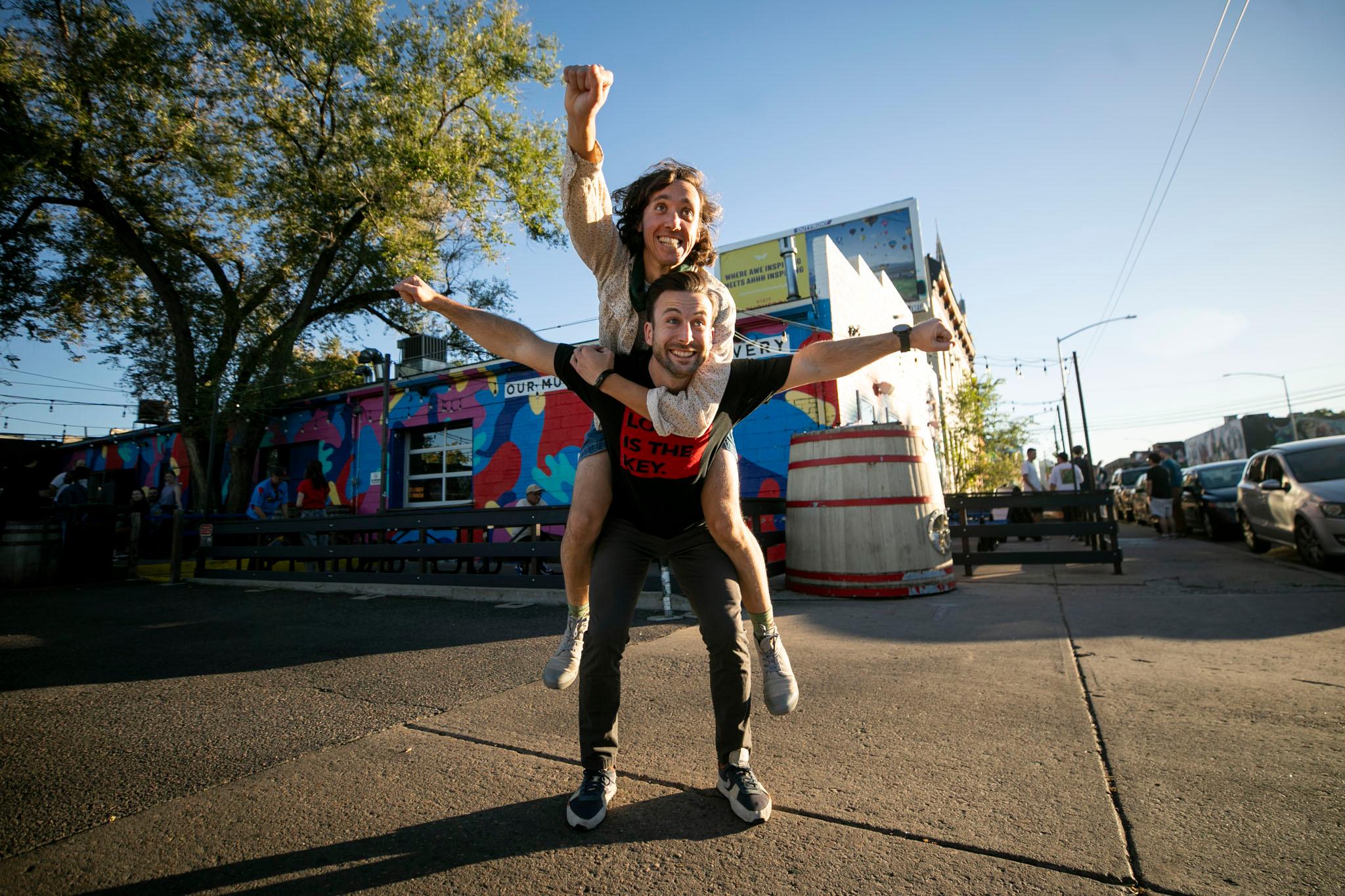 A man in a black t-shirt spreads out his arms like a magnificent eagle as another man, latched onto him as a piggyback rider, raises one arm above his head, as if to say: ONWARD WE GO!