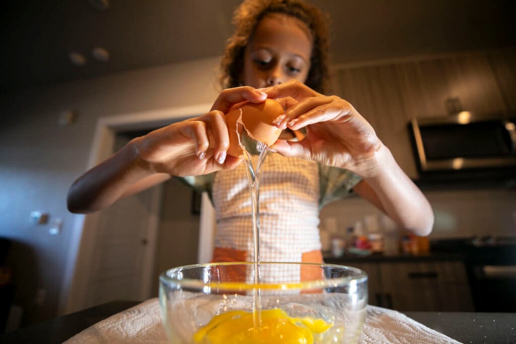 A girl in a plaid apron reaches her hands towards the camera, holding a cracking egg whose whites are oozing out into a bowl beneath it.