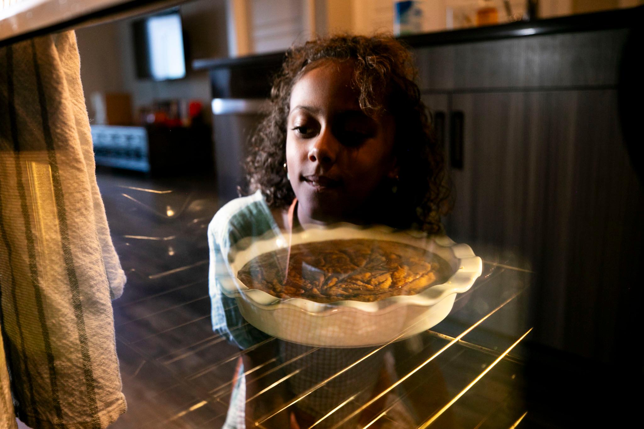 A pie can be seen through the window of an over door; in the oven's reflection, a girl can be seen kneeling and watching it bake.