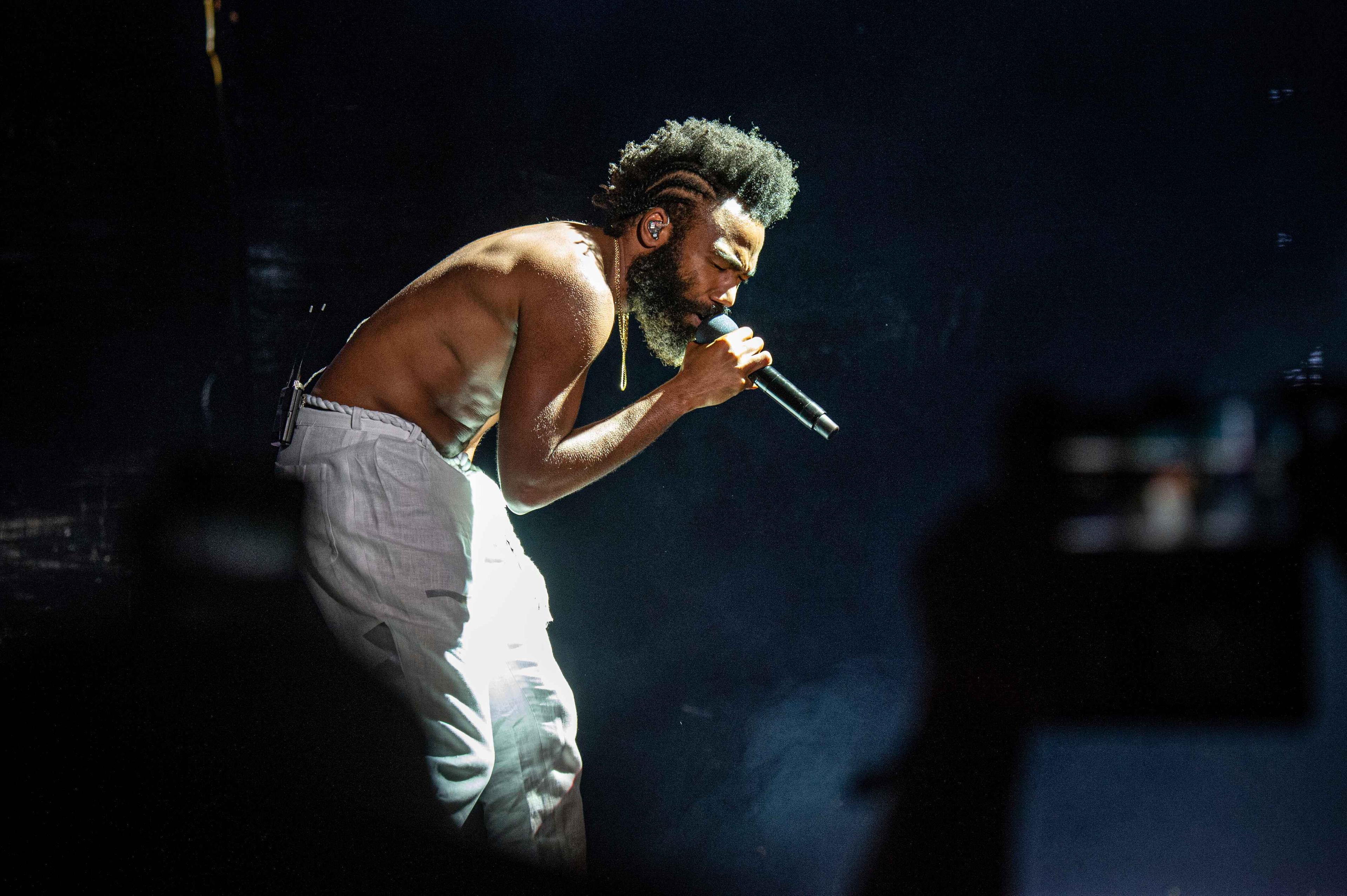 Childish Gambino, dressed in white pants with no shirt, performs at the Bonnaroo Music and Arts Festival on Friday, June 14, 2019, in Manchester, Tenn. He is singing with eyes closed, hunched forward over his microphone.