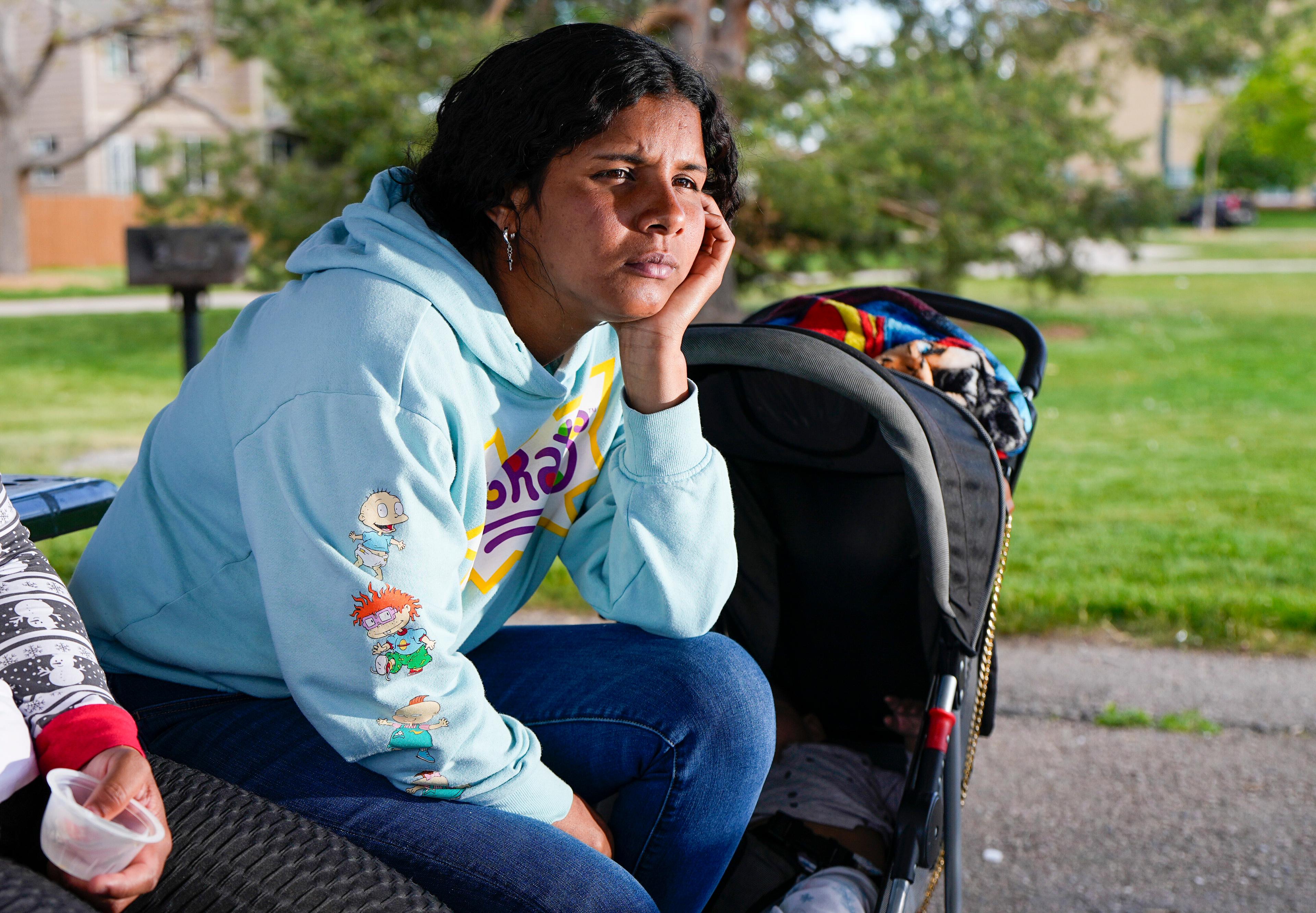 A woman looks past the camera as her face sits on her left hand and her arm is supported by her knee. A baby lays in a stroller to her left.