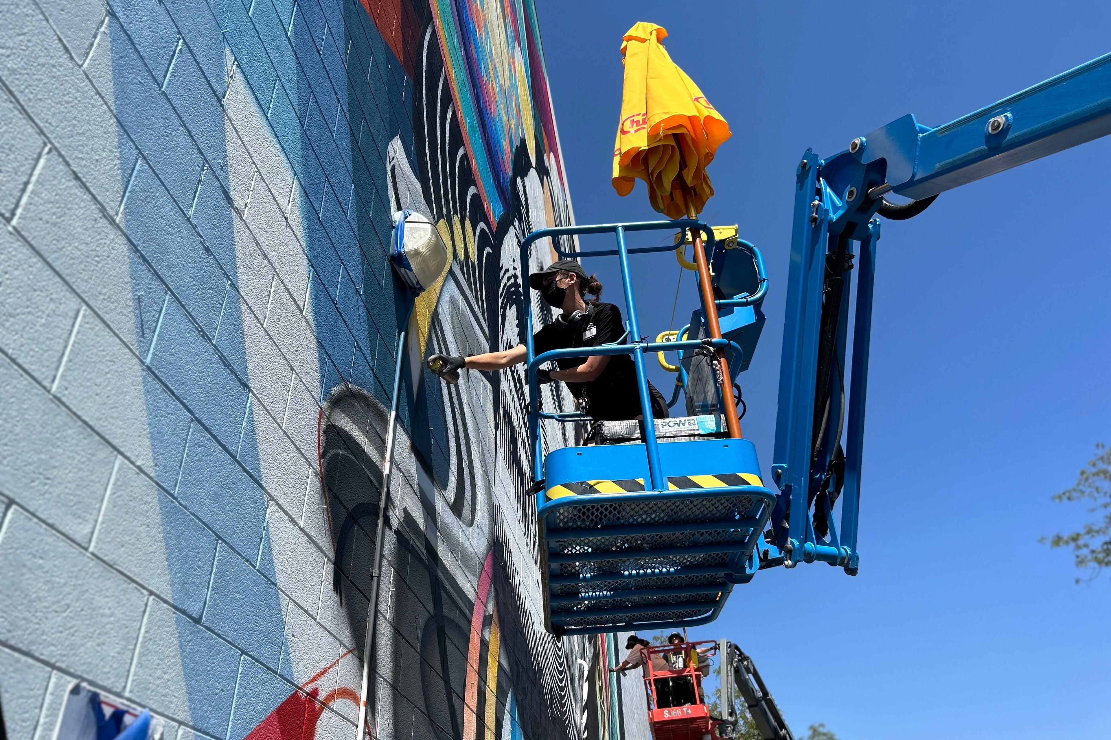 A woman dressed in all black applies spray paint from a telescoping platform to a mural on the side of a building.