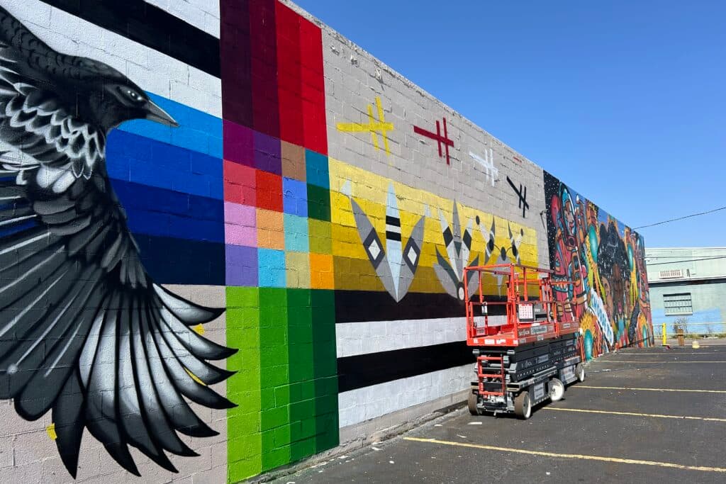 A telesccoping platform sits in front of a colorful mural featuring grey, black and white feathers on a yellow backdrop on the side of a building.