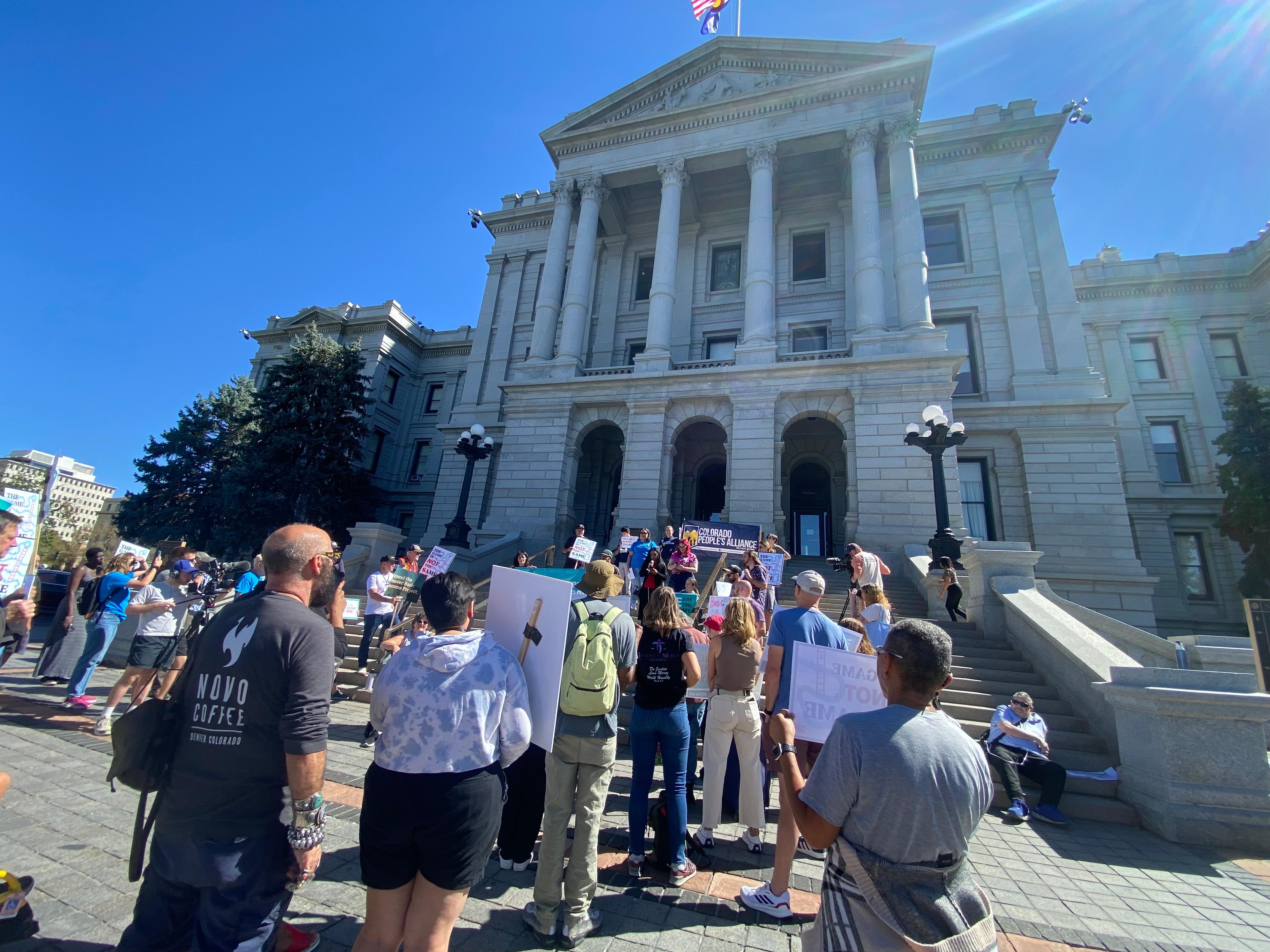 A group of people gather with signs supporting universal basic income in front of the Colorado State Capitol building.