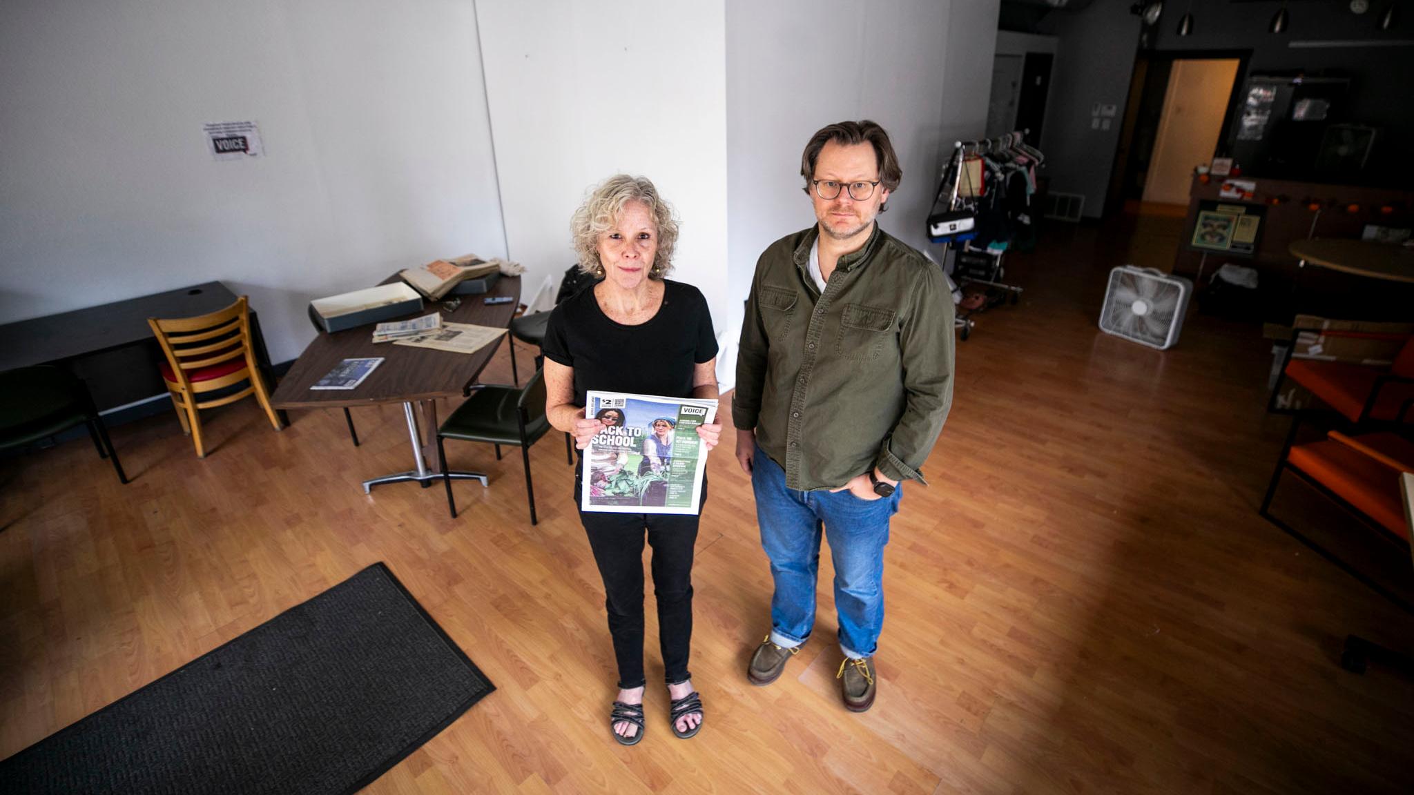 Two people stand in an empty-ish office, looking right into the lens. A woman on the left, with silver hair and a black outfit, holds a newspaper. The man next to her wears glasses and a button-up. They look solemn.