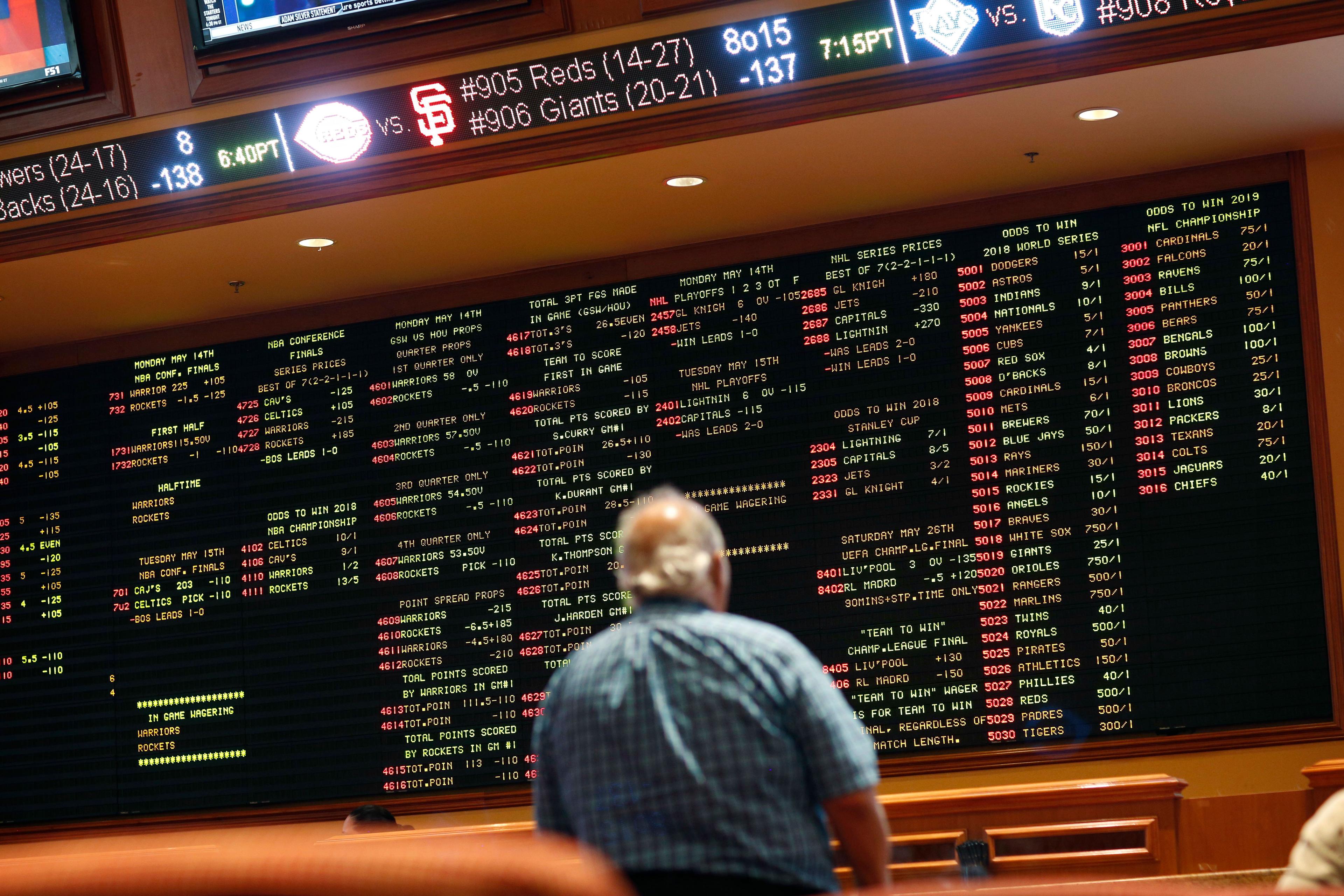 A man stands in front of a digital board displaying sports betting odds.
