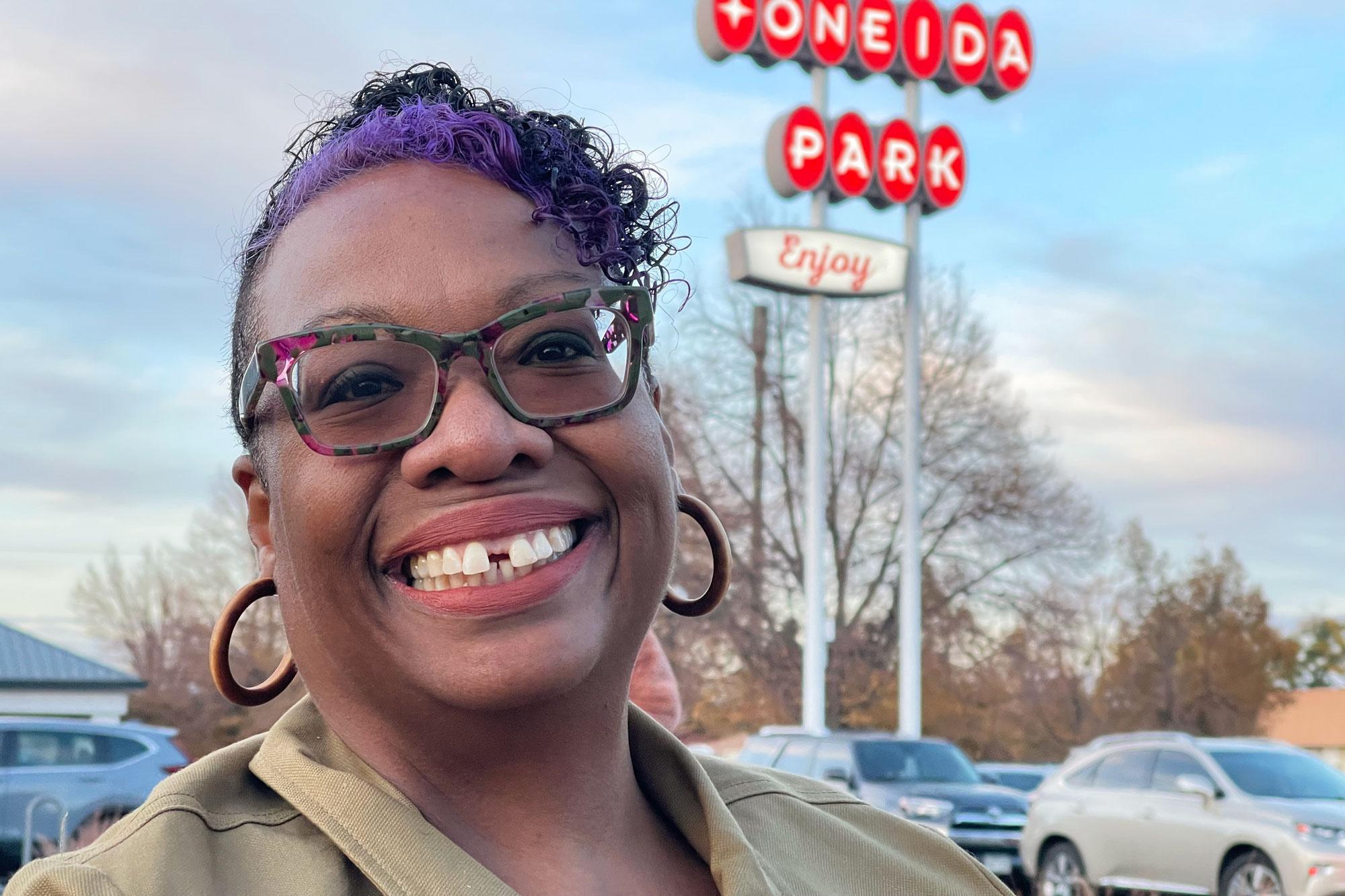 A Black woman with colorful glasses, brown hoop earrings and a purple streak in her hair smiles widely. The retro red sign for the Oneida Park shopping center in Park Hill is seen in the background.