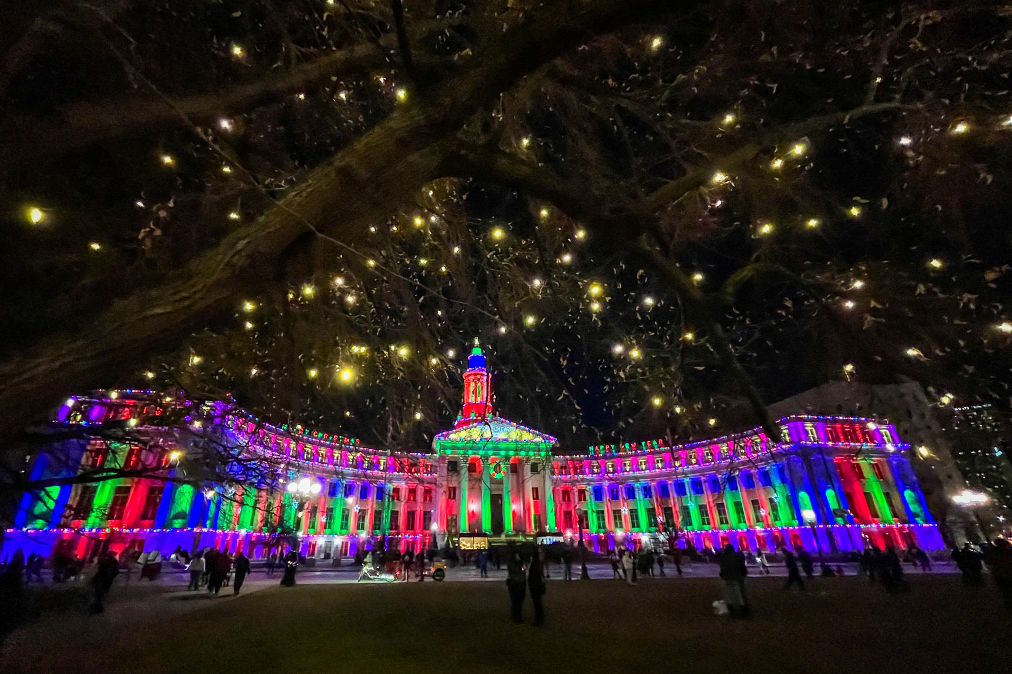 The Denver City and County Building is illuminated with colored lights and framed by a tree hung with yellow and white lights.