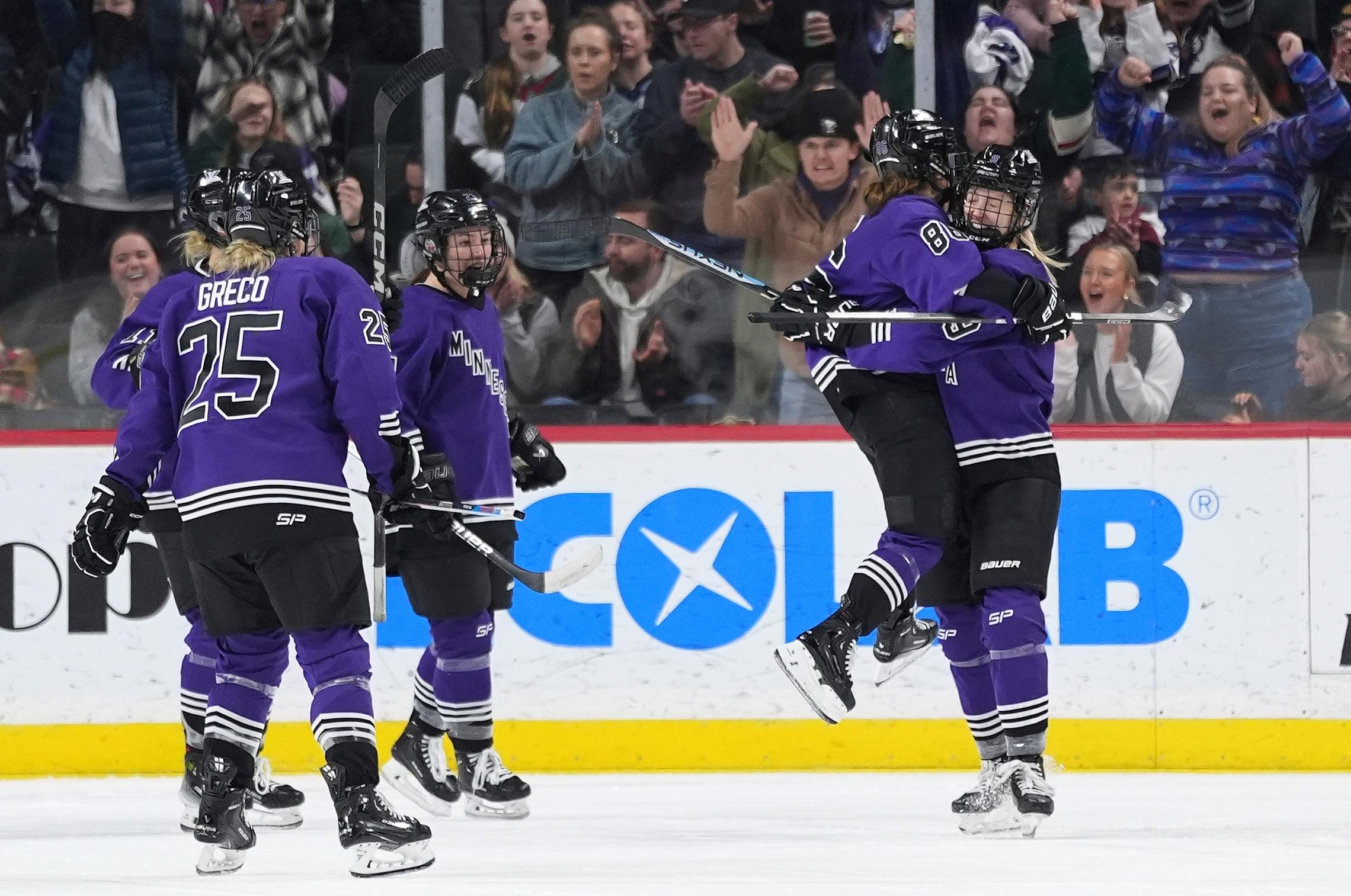 Two women's hockey players embrace after scoring a goal on the righthand side of the image as three more teammates skate over. Fans cheer behind the plexiglass behind them.