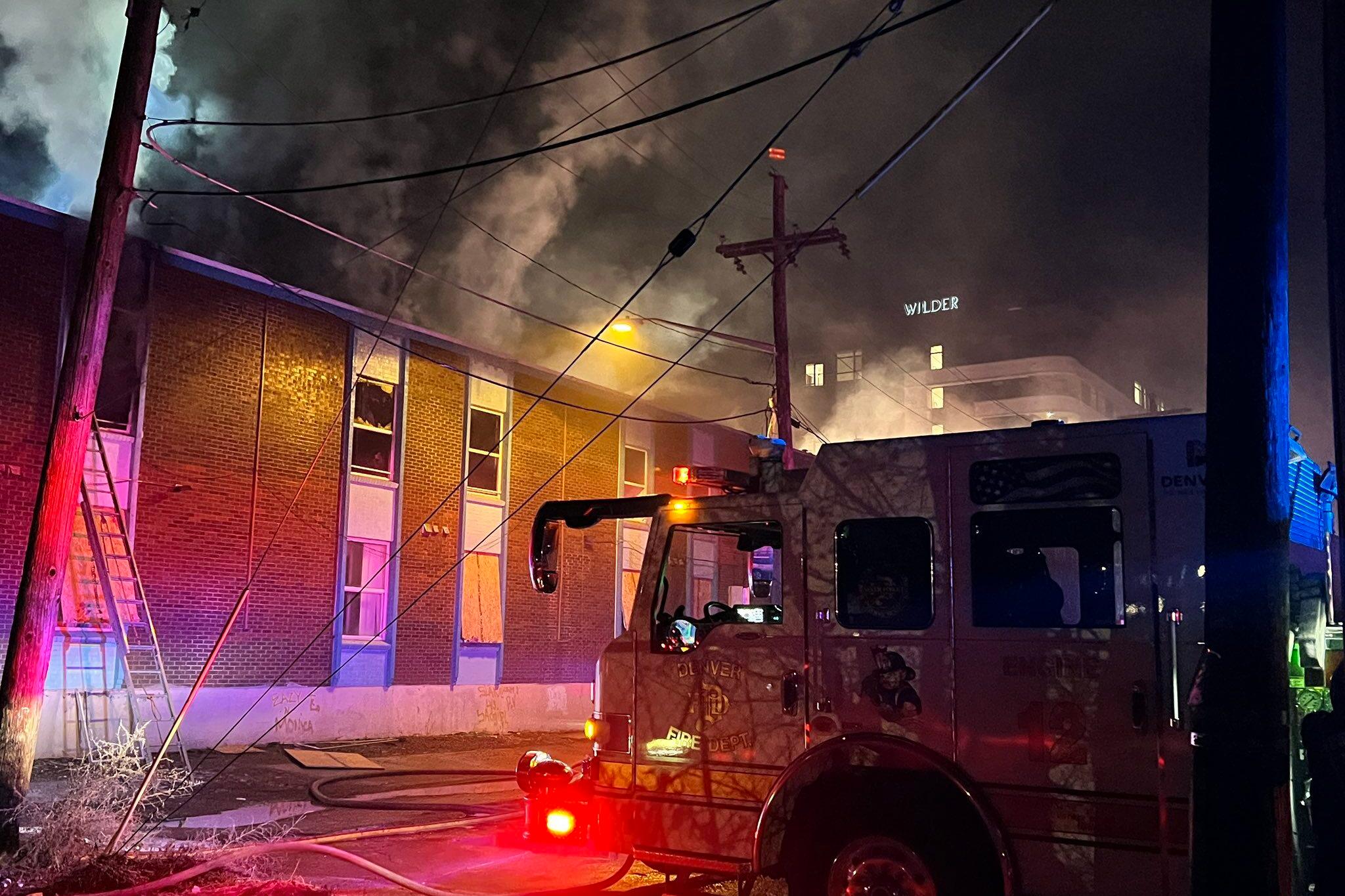 A firetruck is parked near a three-story apartment building that has smoke pouring out from the top. Red and blue emergency lights illuminate the early morning scene.