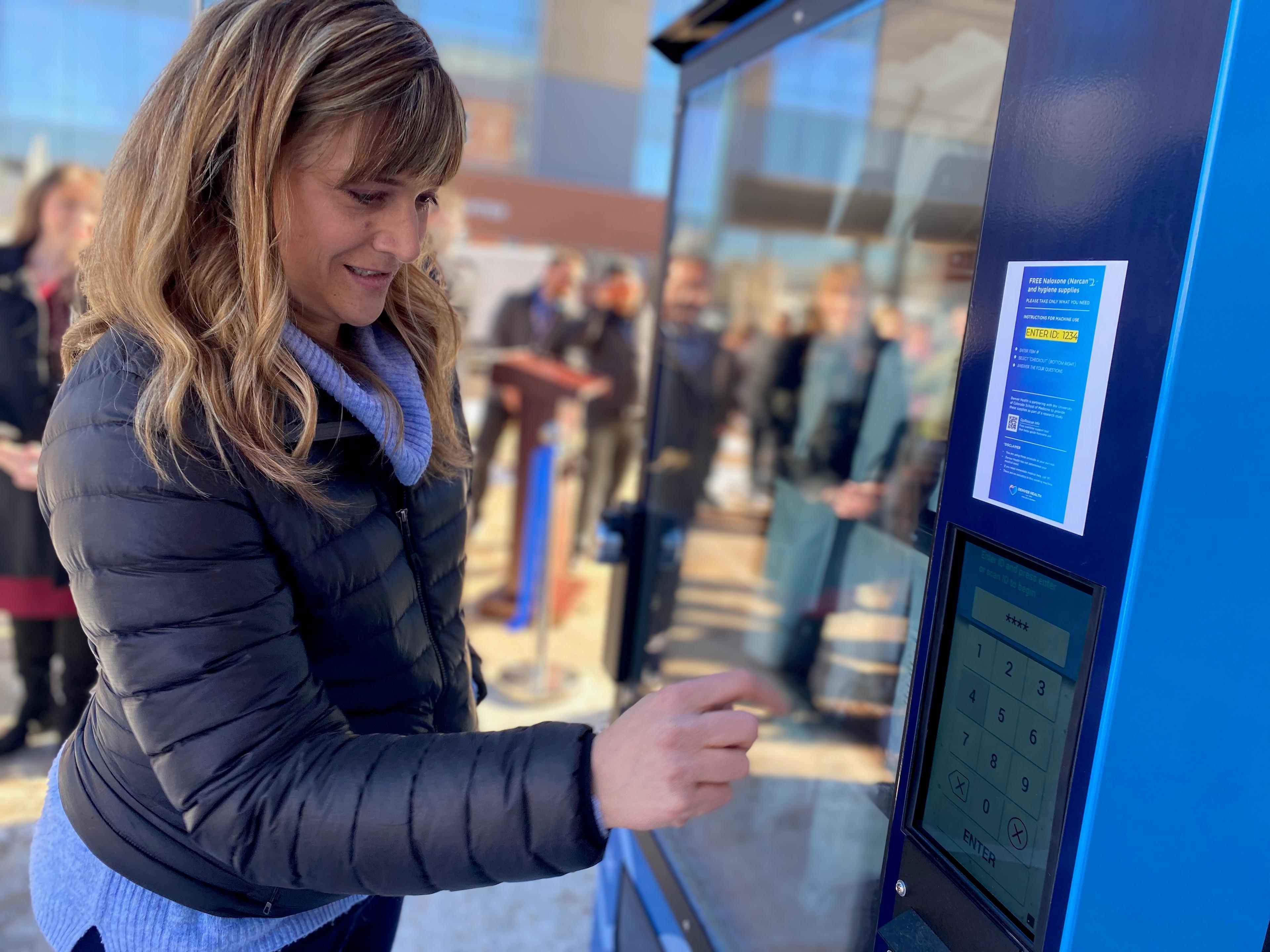 A woman in a puffy jacket tapes a screen on a naloxone vending machine.