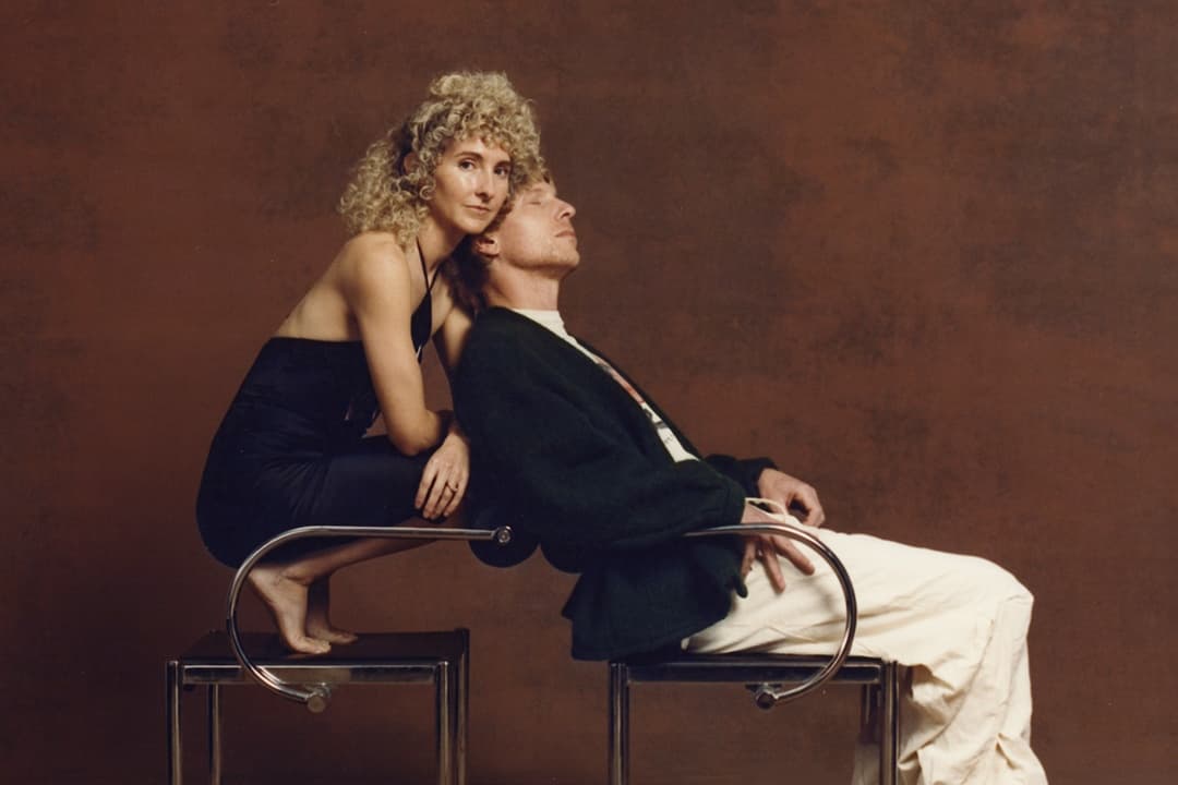 A portrait of two adults. On the left a woman with blond curly hair kneels on a chair, leaning into a man seated in the chair next to her.
