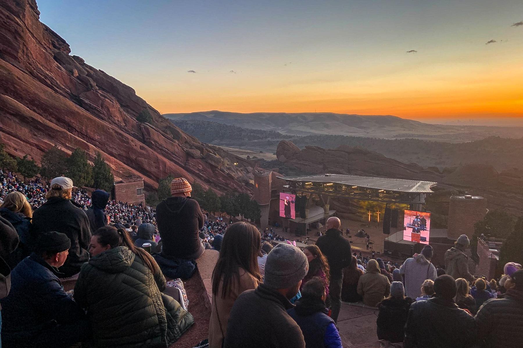 A crowd of people watch the stage at Red Rocks as the sun rises behind it.