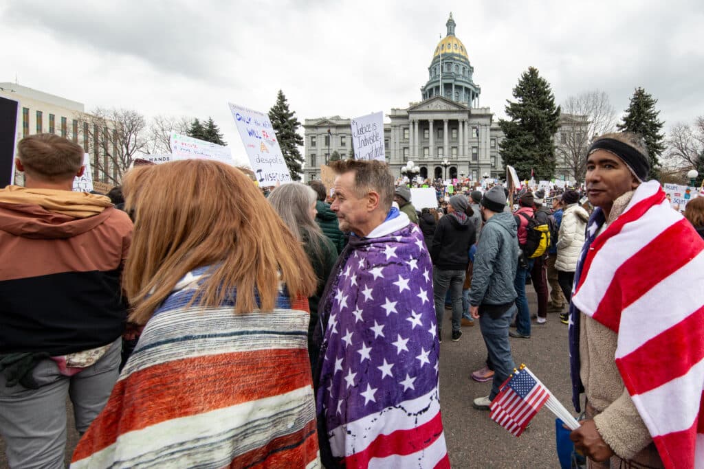 People wrapped in American flags stand in a crowd with Colorado's capitol dome visible behind them.