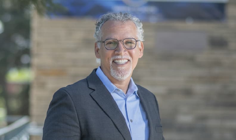 A man with curly grey hair and goatee smiles in front of a brick building. he's wearing a classic suit with no tie and has glasses on.