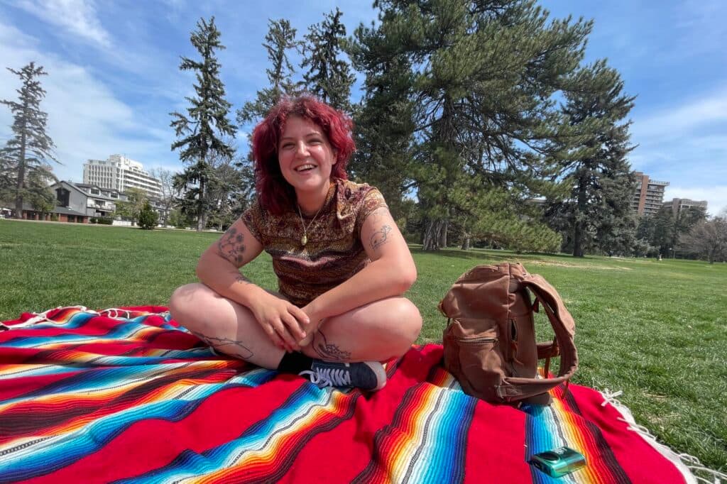 A young woman sits on a colorful striped blanket in the middle of a green park. She has purply-red hair and is grinning.