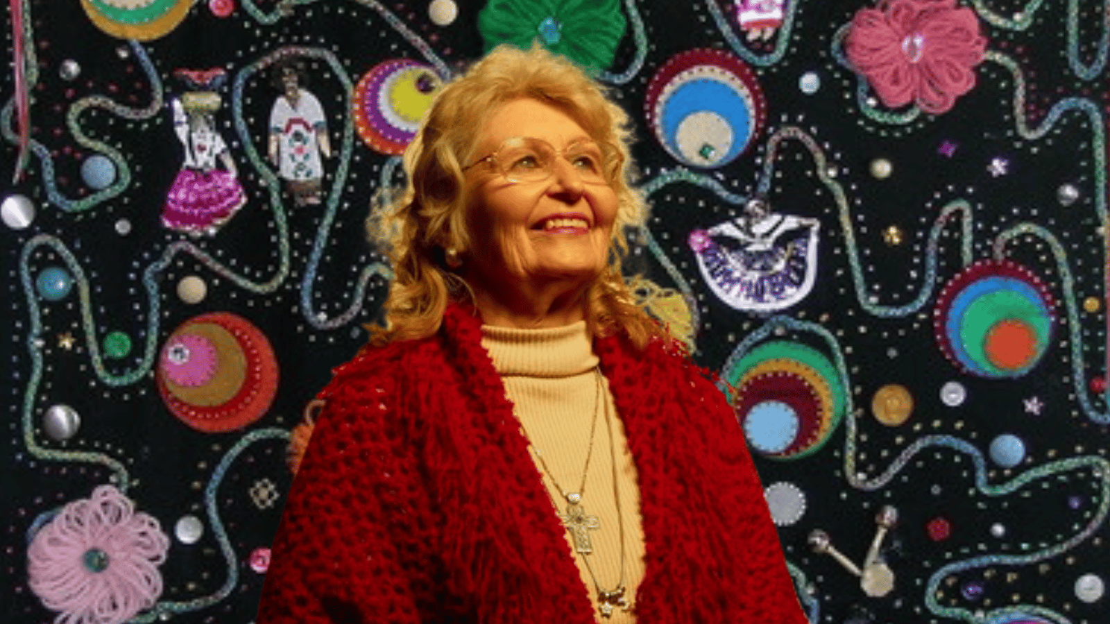An older woman with glasses an blonde-ish hair smiles. She wears a cross necklace and chunky red cardigan over a yellow turtleneck. She has been photoshopped against a background of her folk art, depicting traditional dolls, colorful circles and flowers.
