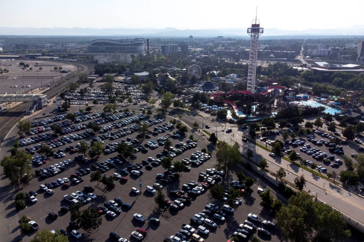 An aerial view of Elitch Gardens, its white view tower breaking the mountain-lined horizon, and a big, full parking lot.
