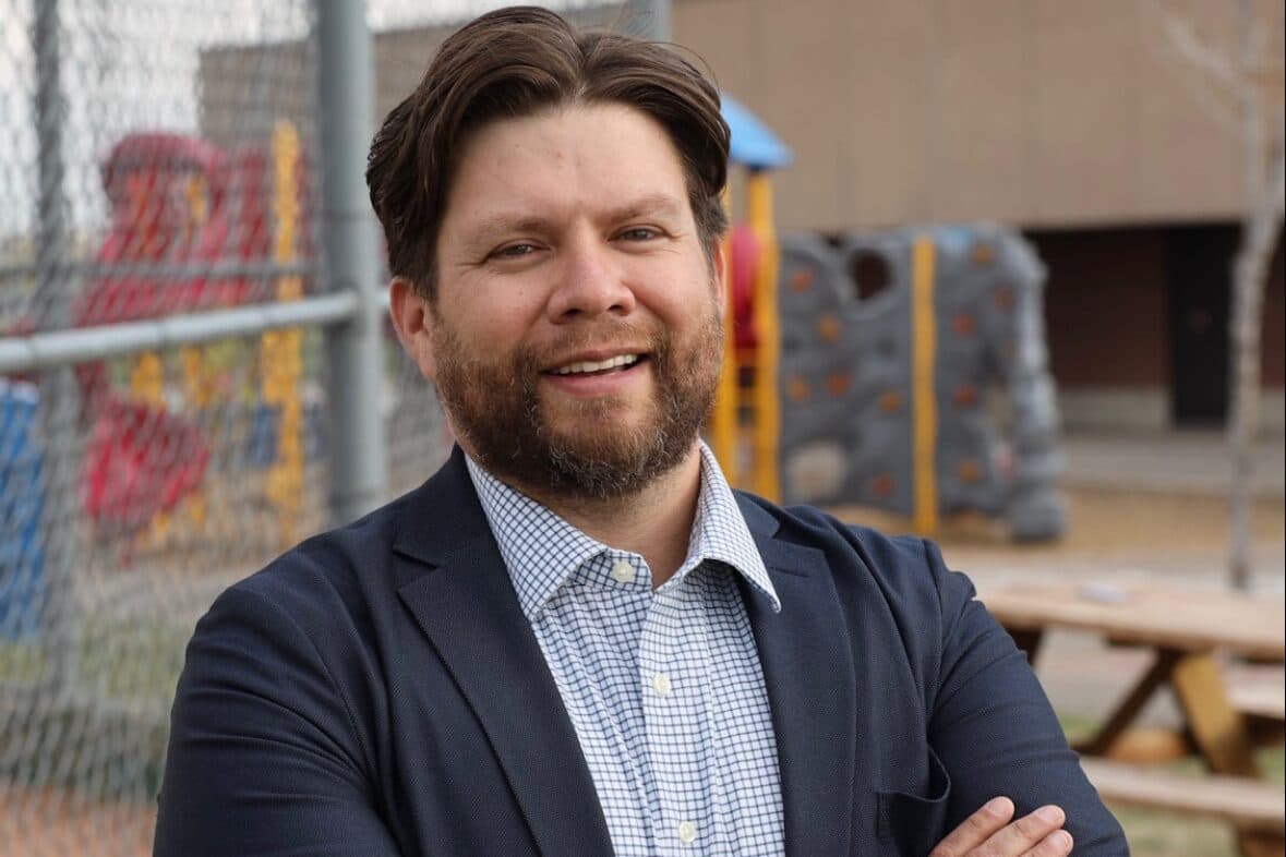 A man with brunette hair and a beard smiles at the camera with his arms crossed. He wears a blue button-up shirt and navy blazer.