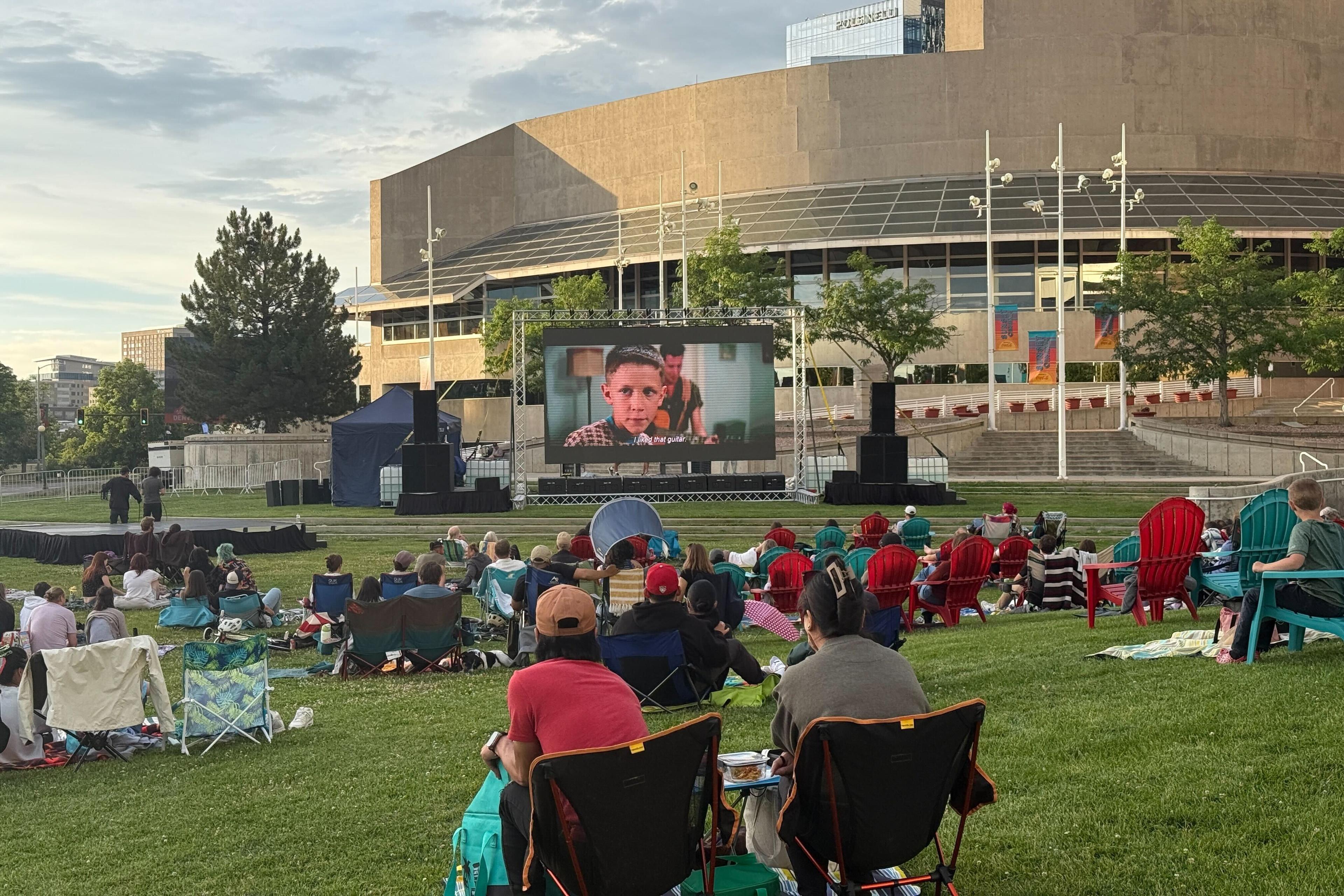 A crowd of people sit on lawn chairs and blankets on a lawn at sunset. A large screen shows "Forrest Gump," and Denver's art complex sits in the background.