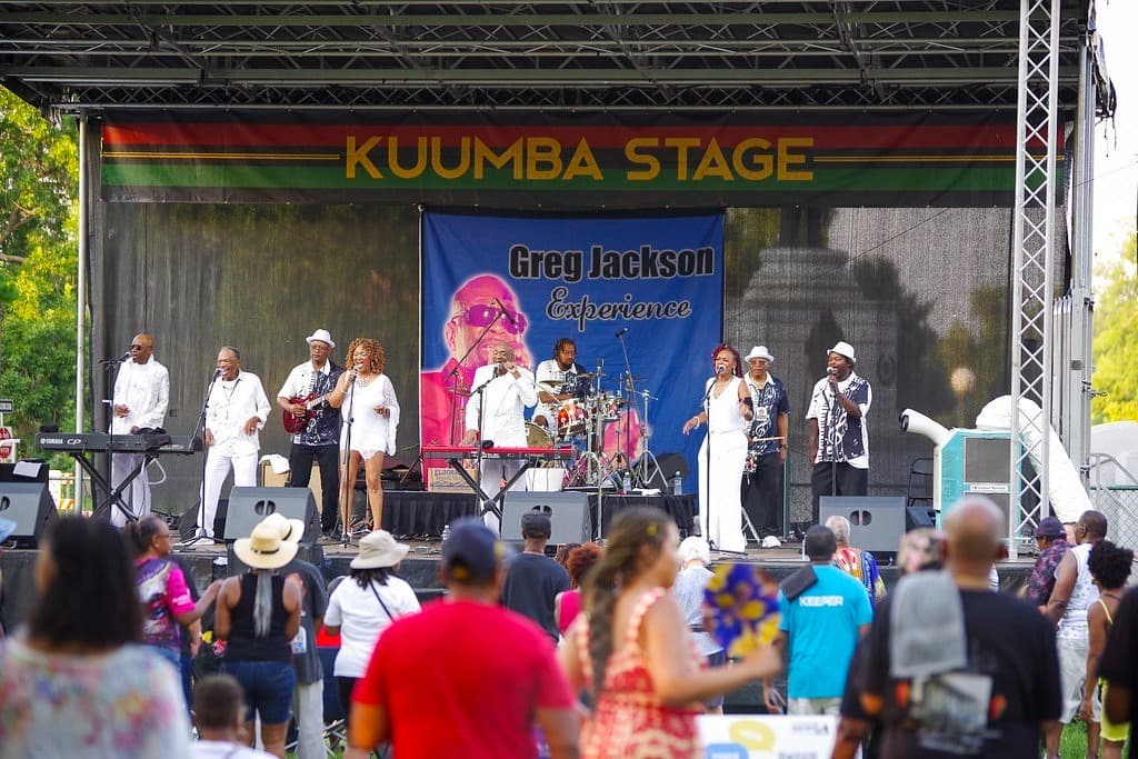 Photo shows a band performing on a stage. The performers are mostly wearing white outfits. Across the top of the stage is a striped banner that reads "Kuumba Stage." There is an audience standing in the foreground.
