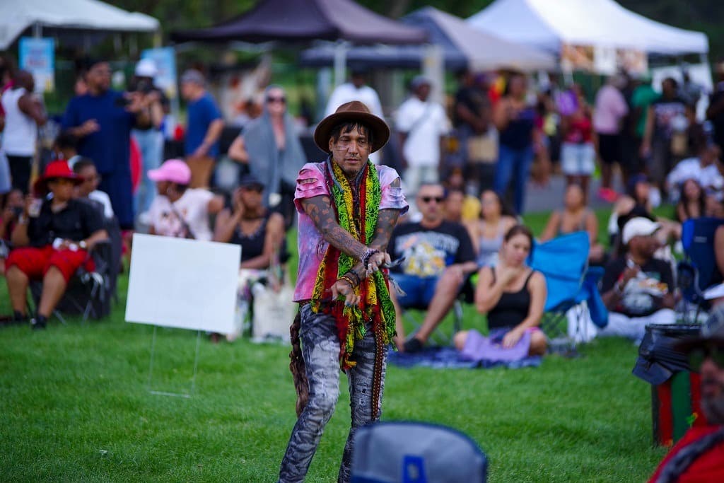Photo shows festival tents and crowd in a blurred background. In the center of the photo is a man with a colorful scarf and tee shirt with his arms stretched and crossed in front of him. He is wearing a fedora.