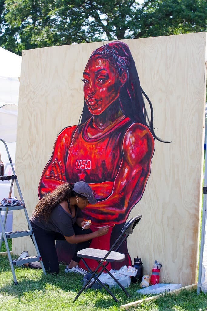 Photo shows an artist crouched on the ground painting the image of an Olympic athlete in red.