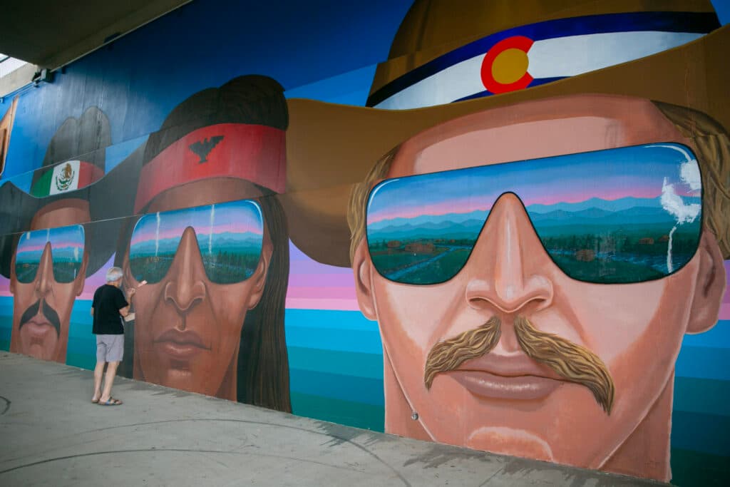 Emanuel Martinezstands in front of three large, painted faces on the wall, holding a paint brush. The dark-skinned figure on the far left wears a cowboy hat with a Mexican flag hatband, the dark-skinned figure in the middle wears a red headband with a thunderbird, and the light-skinned figure on the right wears a cowboy hat with a Colorado flag hatband.