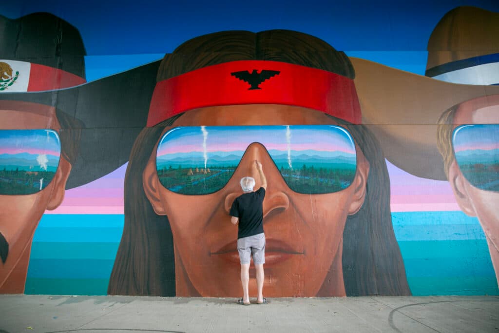 Emanuel Martinez uses light tan paint to touch up the nose of a large head painted on the wall. The figure is an indigenous man in a red headband bearing a thunderbird. The man's glasses reflect a tipi village at the confluence of the South Platte River and Cherry Creek.