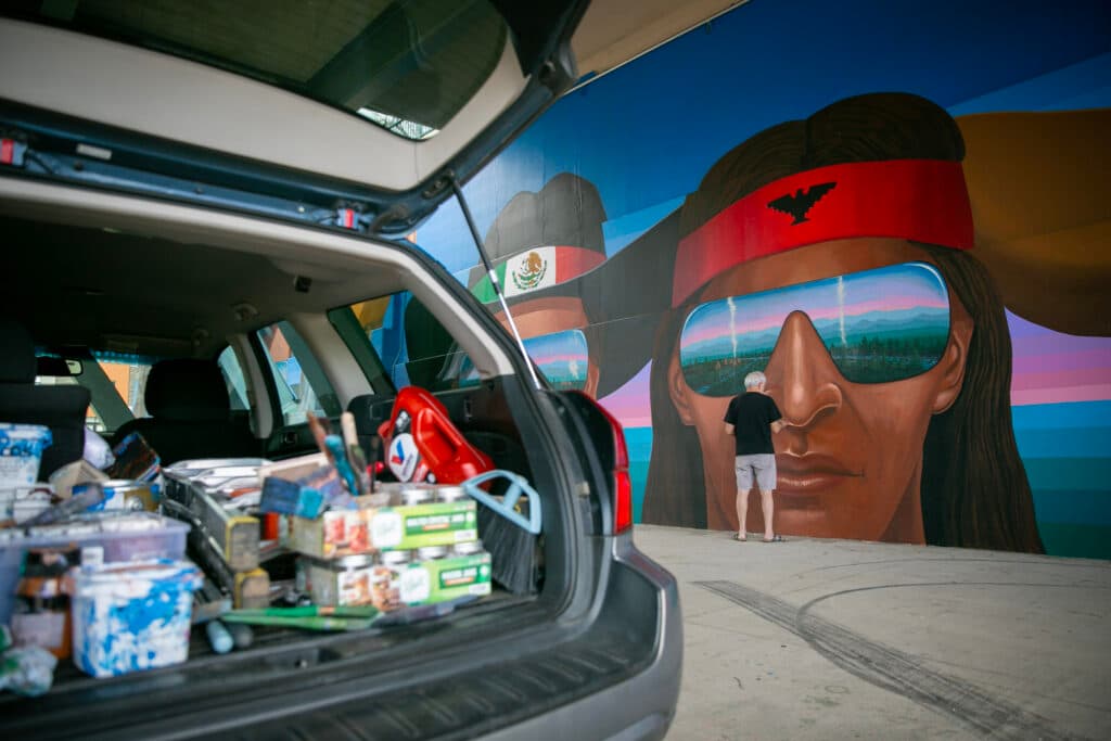 Emanuel Martinez uses light tan paint to touch up the nose of a large head painted on the wall. In the foreground, a hatchback Subaru is open, reveling a trrunk of paint supplies, jars, a broom head, a ladder, and more.